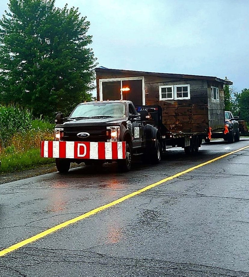 Un camion Ford transporte une petite maison en bois gris foncé avec des fenêtres blanches, sur une route mouillée. La maison a une porte et deux fenêtres visibles, avec des arbres verts en arrière-plan.