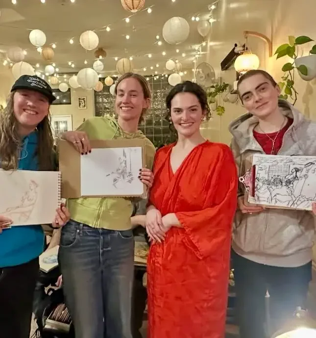 Four participants at Drink N Draw at Bat Haus in Brooklyn holding their sketchbooks with figure drawings, standing in the warmly lit event space with string lights overhead.