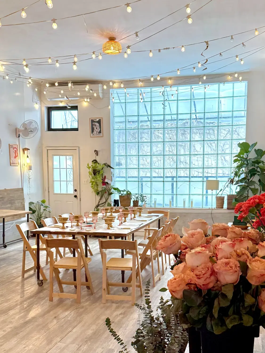 Bat Haus Brooklyn event space set with a long table, peach rose centerpieces, string lights, and a glass-block window in Williamsburg.