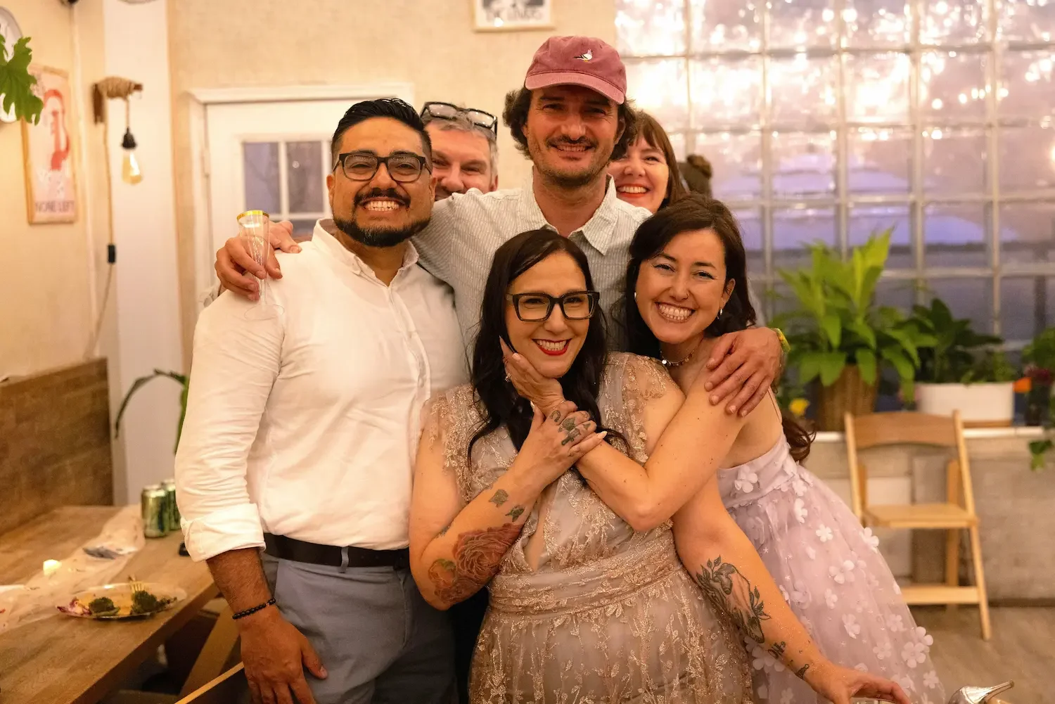 Bride in an embroidered blush gown embraced by smiling friends in front of the glass-block window at Bat Haus, an intimate wedding venue in Brooklyn