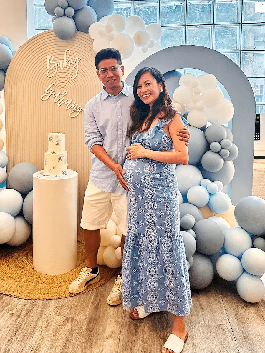 Expecting couple smiling beside a blue balloon arch and tiered cake at a baby shower at Bat Haus in Williamsburg