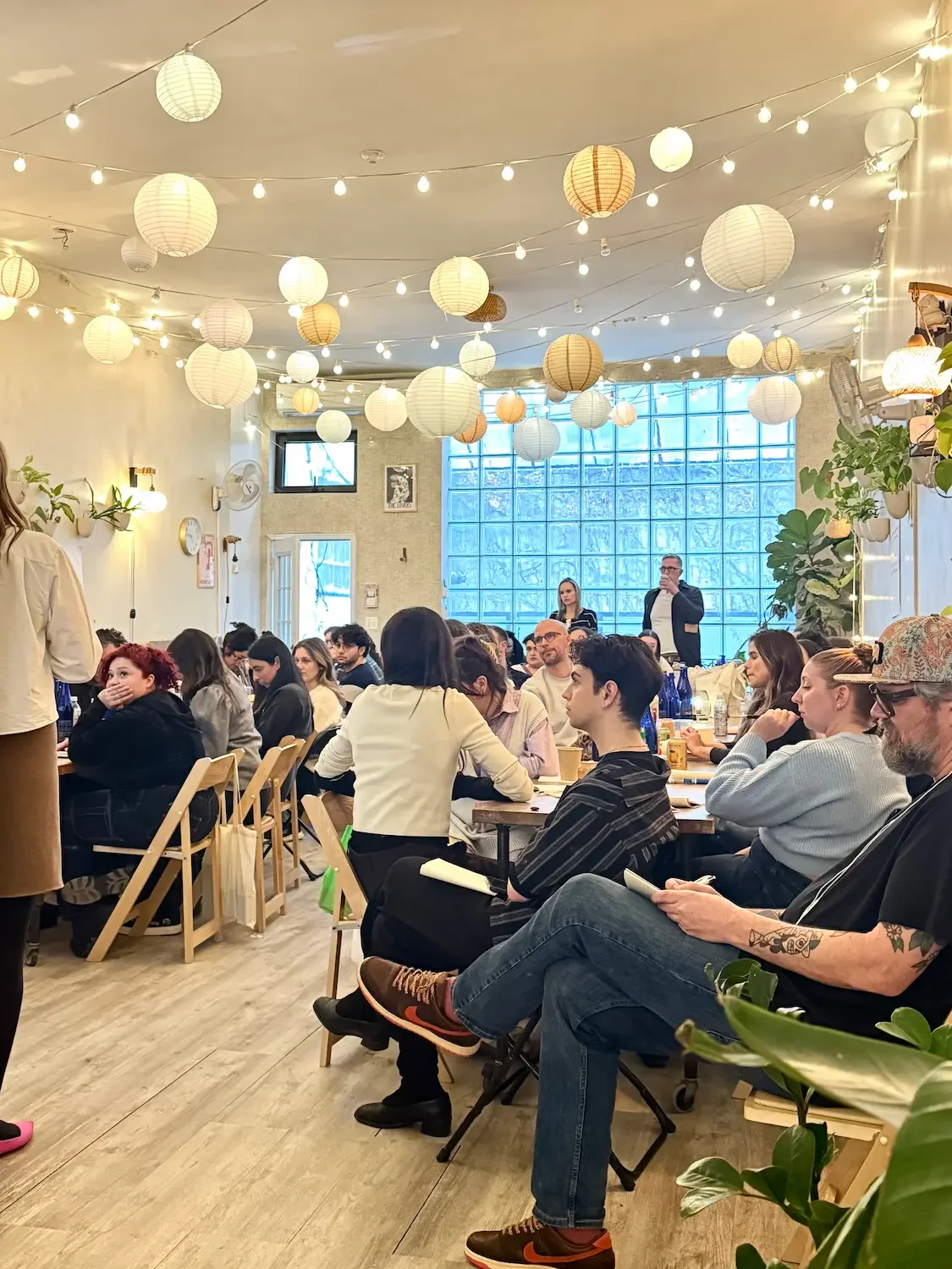 Team seated at long tables during a corporate offsite at Bat Haus Brooklyn event space, with paper lanterns, string lights, and a glass-block window in Williamsburg.