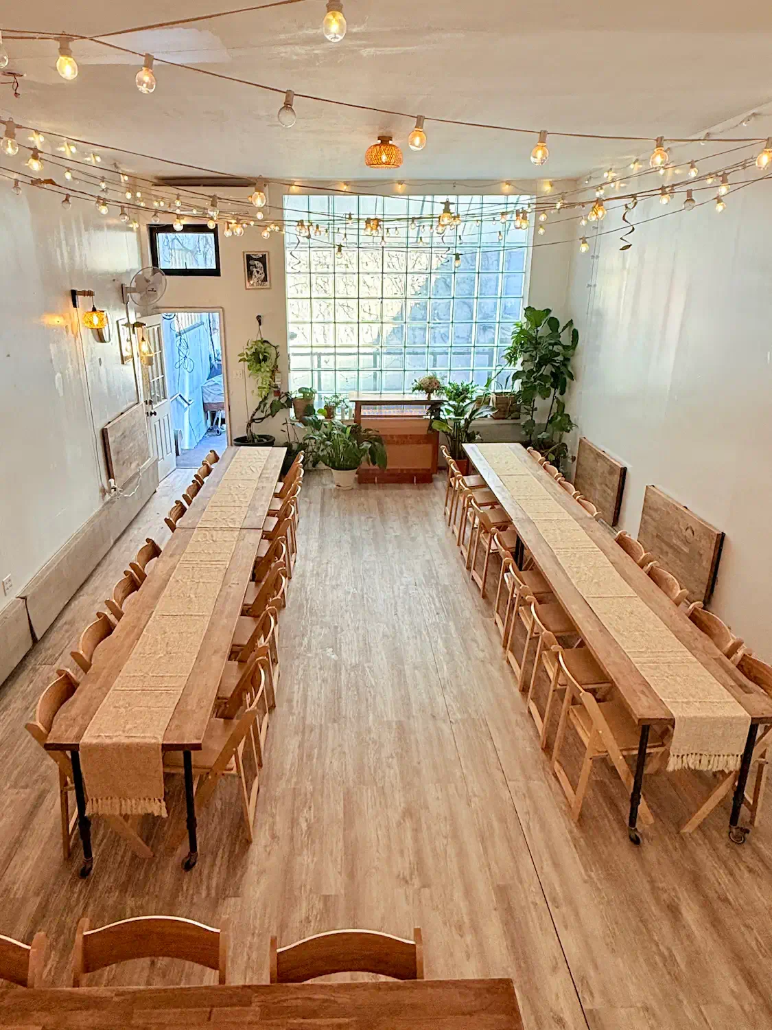 Light-filled event space in Williamsburg Brooklyn with two long wooden tables, string lights, glass block window, and access to a private backyard, set up for a wedding, dinner party, or corporate offsite at Bat Haus.