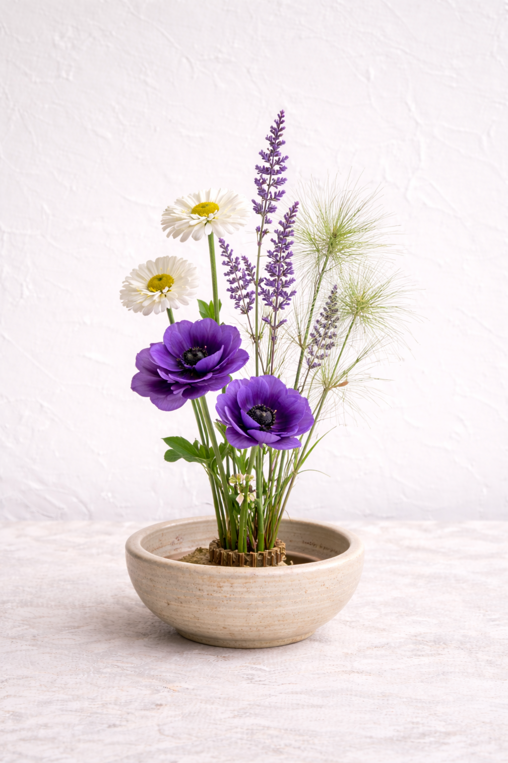 A sculptural Japanese ikebana floral arrangement with coral dahlias, burgundy ranunculus, blush roses, and airy seasonal accents, arranged in a handmade ceramic bowl. The composition embraces asymmetry and negative space, offering a refined, modern i