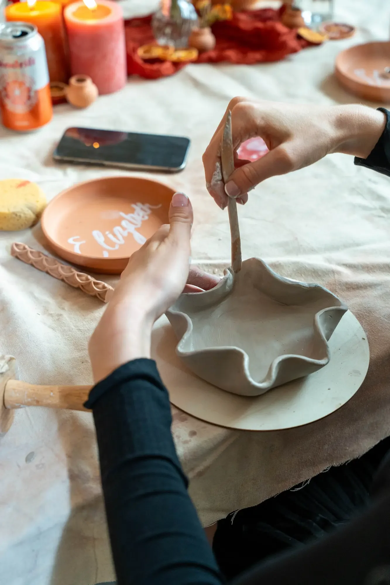 Guest's hands sculpting a ruffled clay dish with a wooden tool at a Brooklyn pottery party, beside a personalized terracotta name plate.