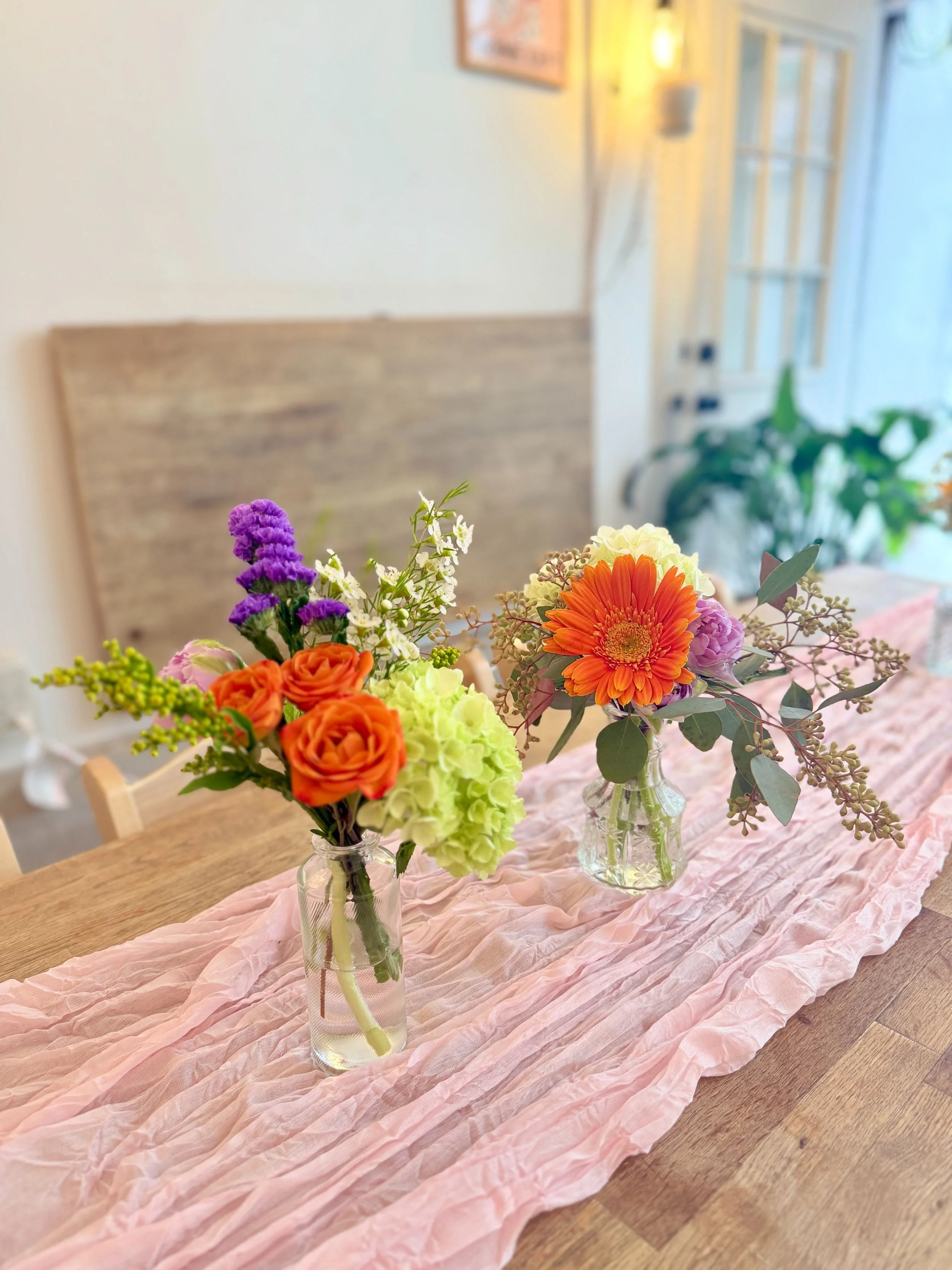 Two coordinating mini vases—orange roses with green hydrangea and a gerbera mix—styled on a blush runner for a welcoming, photo-ready table.