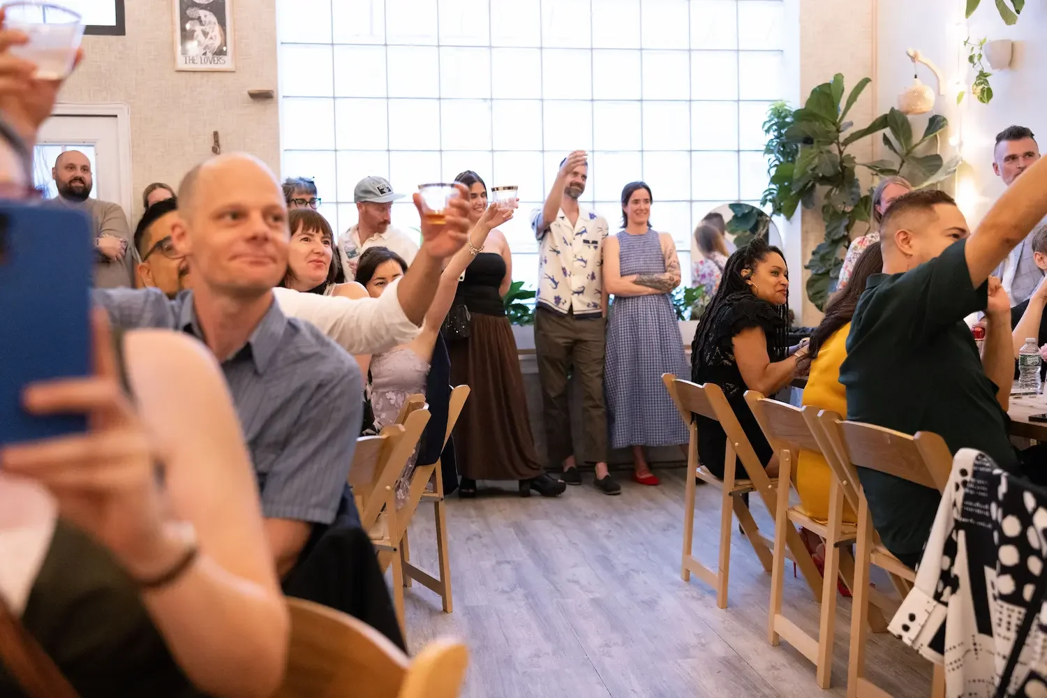 Forty wedding guests raising glasses in a toast under the large glass-block window at Bat Haus, an intimate wedding venue in Williamsburg, Brooklyn