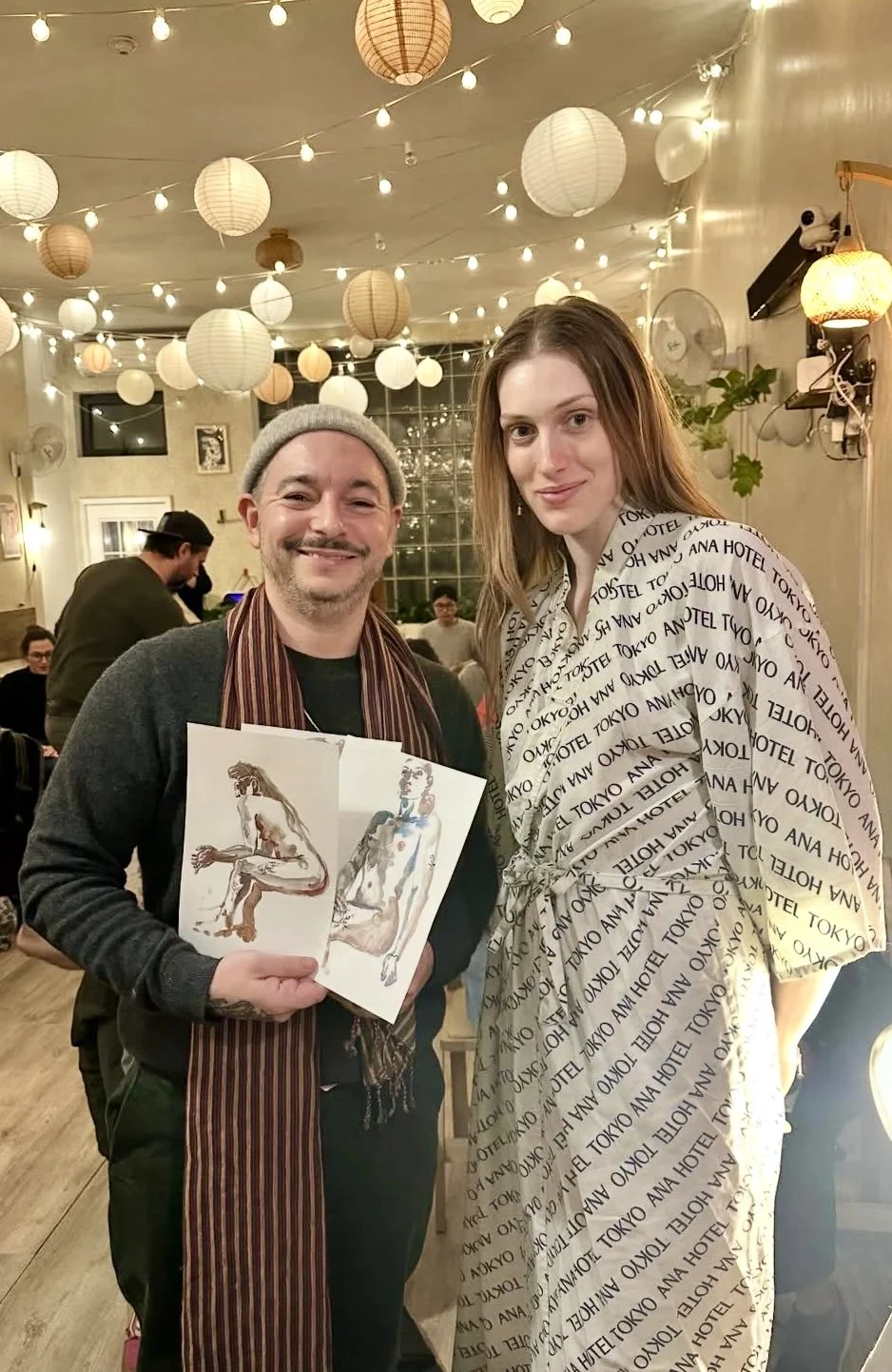 Regular Drink n Draw participant at Bat Haus Brooklyn displaying his finished figure drawing while posing with the live model after the session, a welcoming weekly art event and drawing class in Brooklyn