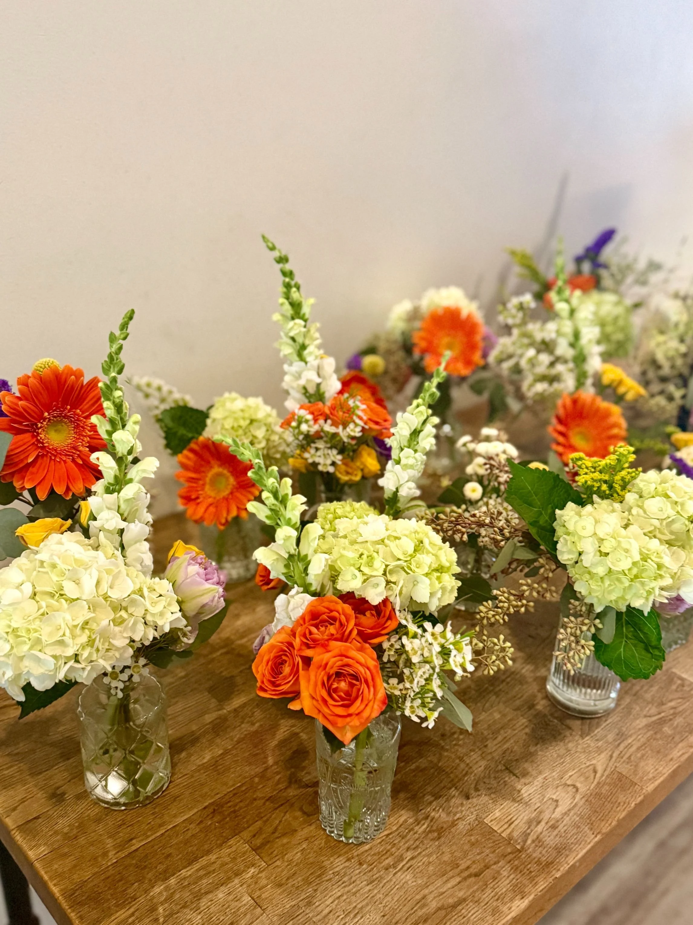 A mix of small glass vases filled with hydrangea, snapdragons, roses, gerbera, and seasonal filler—ready to line Bat Haus’s long tables.