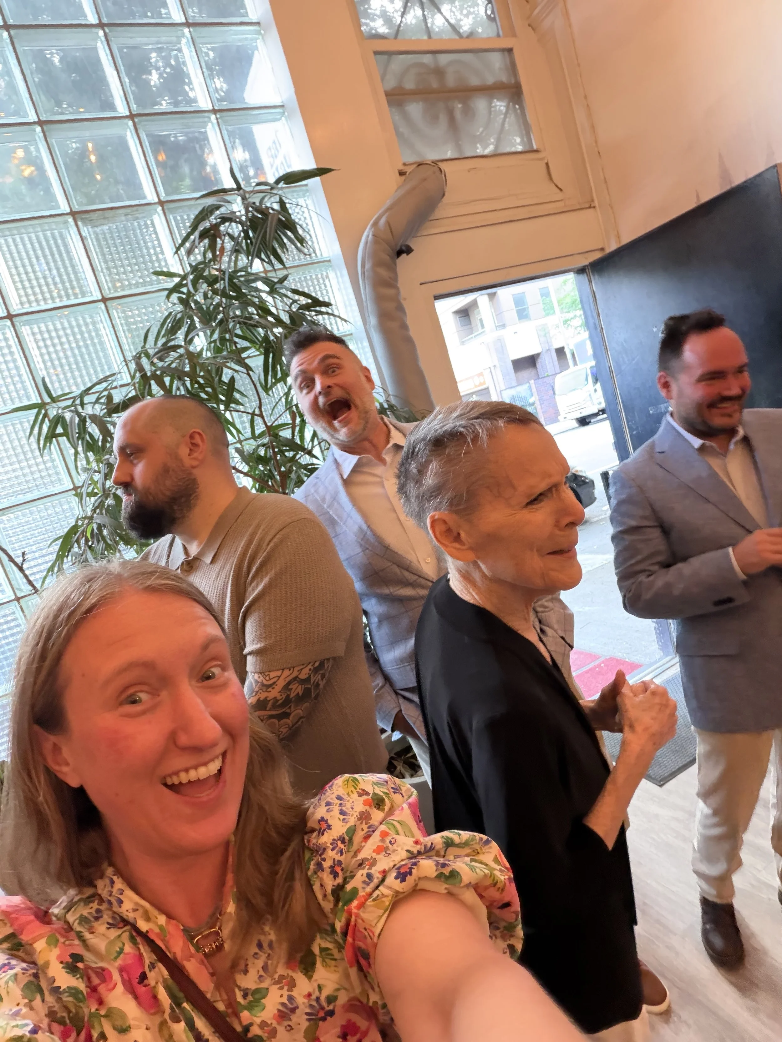 Wedding guests laughing in a candid moment by the sunlit entrance and glass-block window at Bat Haus on Grand Street in Williamsburg, Brooklyn