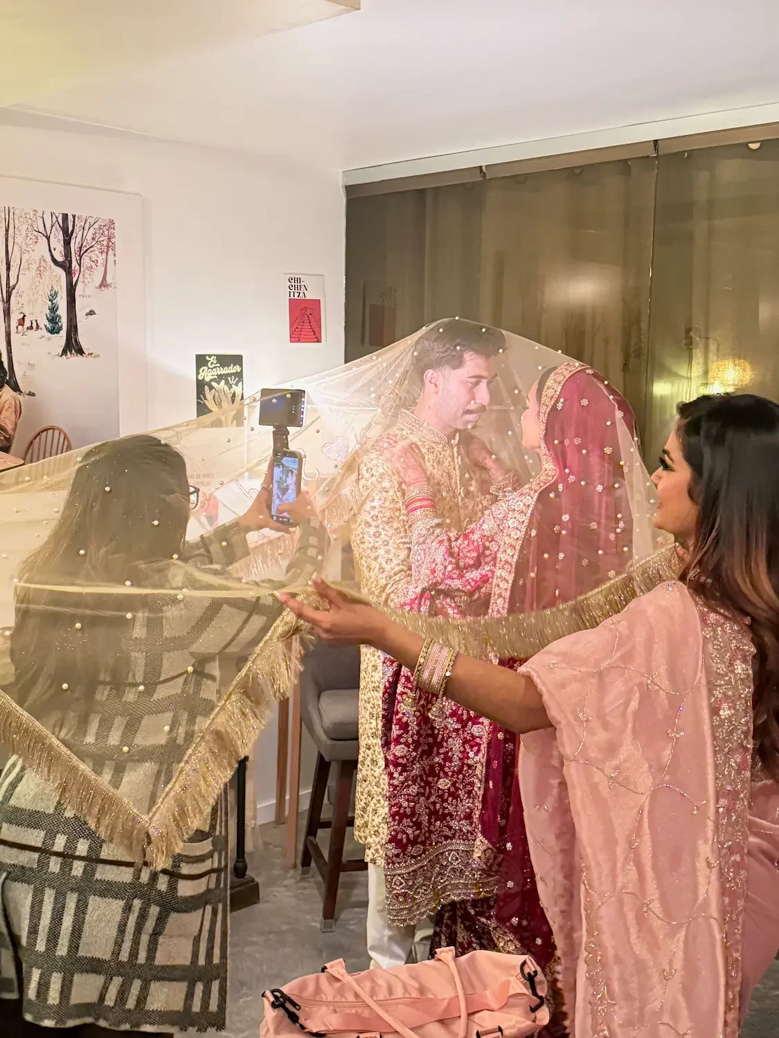 Bride in an embroidered deep pink lehenga and groom in a gold sherwani share a moment beneath a sheer pearl-studded veil held by friends at a Nikah celebration at Bat Haus in Brooklyn