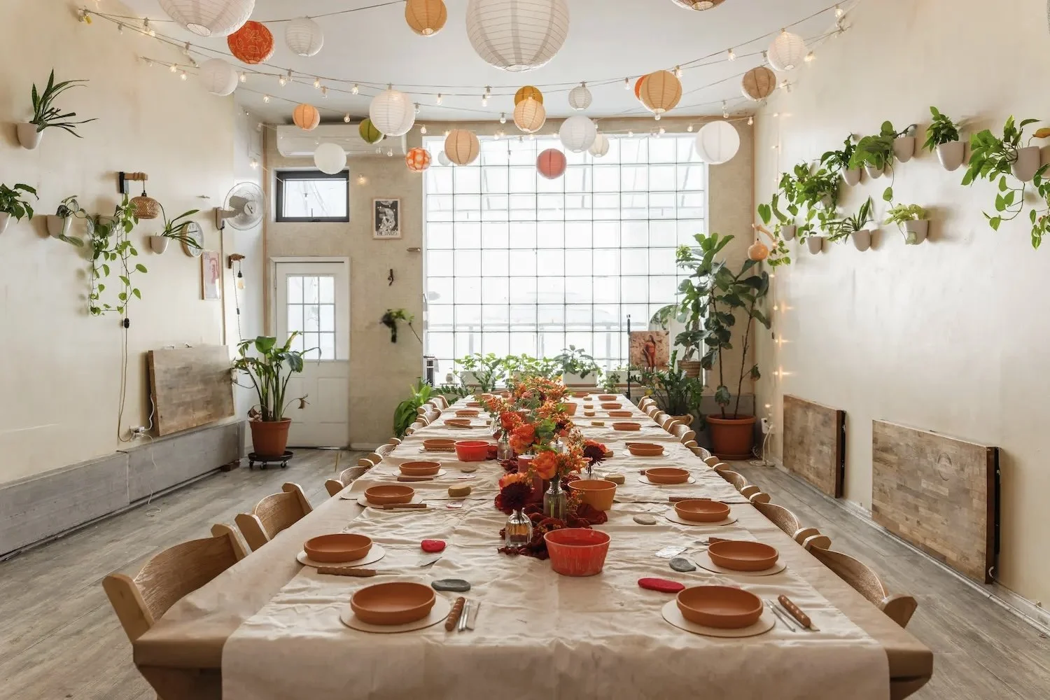 Long table set for a brand event pottery workshop at Bat Haus in Williamsburg, Brooklyn, with terracotta pottery pieces, fall florals, paper lanterns, and a sunlit factory window.