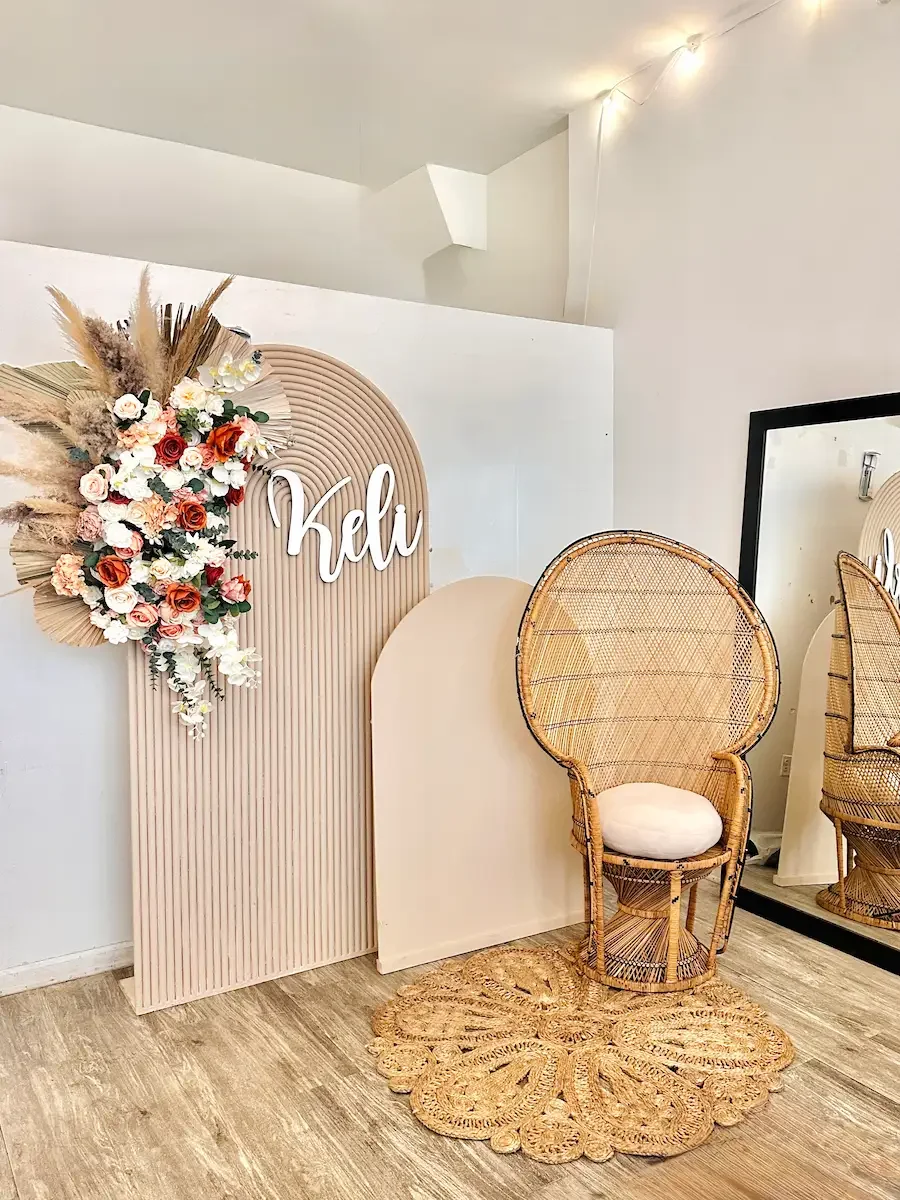 Styled event backdrop corner at Bat Haus Brooklyn featuring neutral arch panels, floral arrangement, custom name sign, and rattan chair for photo moments.