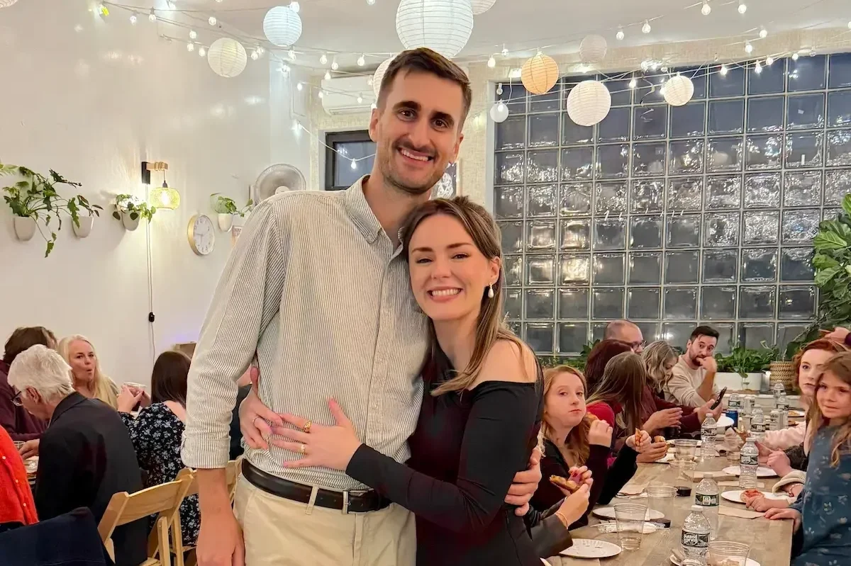 A newly engaged couple embracing in front of the not-see-through glass-block window at their engagement party at Bat Haus in Brooklyn.