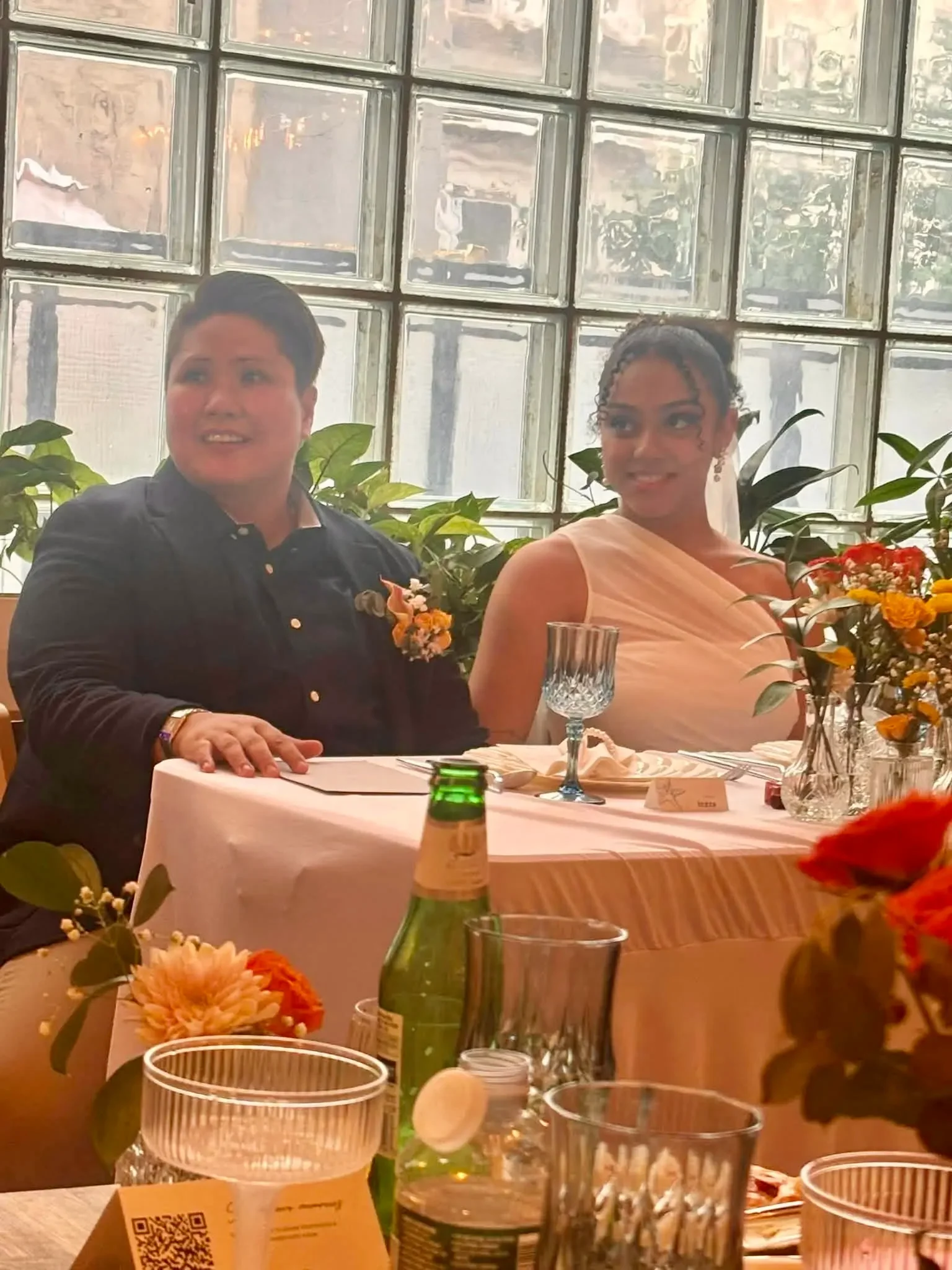 Newlywed couple seated at their sweetheart table with orange florals in front of the glass-block window at Bat Haus, an LGBTQ+-welcoming wedding venue in Brooklyn