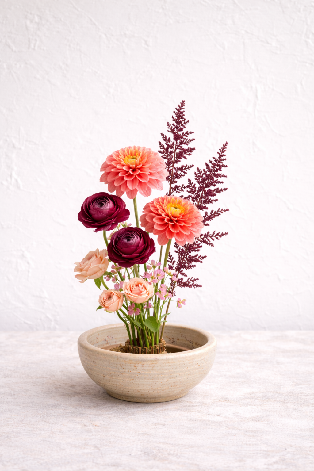 A minimalist Japanese ikebana arrangement featuring coral dahlias, deep burgundy ranunculus, blush garden roses, and delicate seasonal florals, artfully composed in a handcrafted ceramic bowl. Designed with asymmetrical balance and natural movement, 