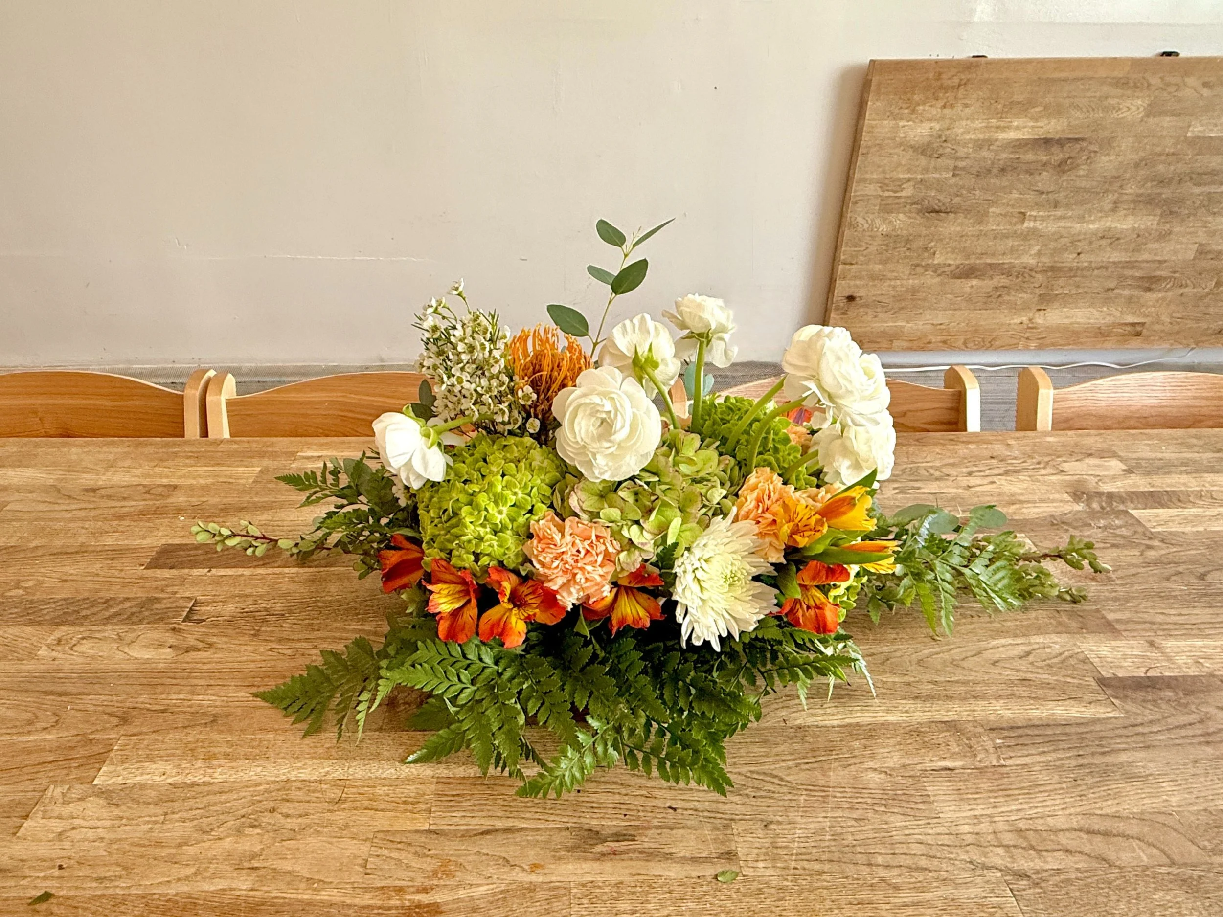 Modern Grove — Cream, Green & Amber
Centered whites (ranunculus, mums) anchored by green hydrangea and warm amber accents, finished with airy fern. Compact height with graceful length—clean, modern lines without blocking faces.
Vibe: polished, balan