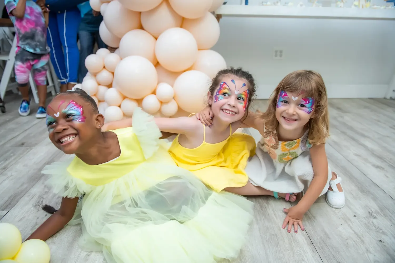 Three girls with butterfly face paint smiling by a balloon arch at a kids' birthday party at Bat Haus in Brooklyn.