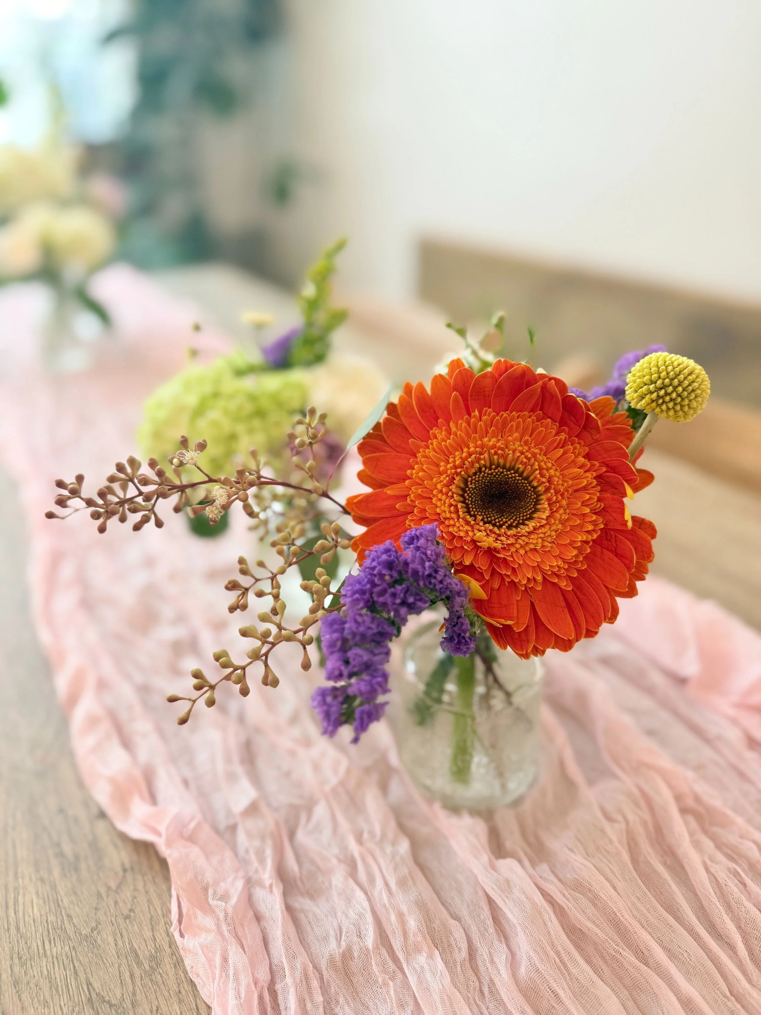 Close-up of a mini vase with orange gerbera, purple statice, and textural greens on a blush runner—small scale, big impact.