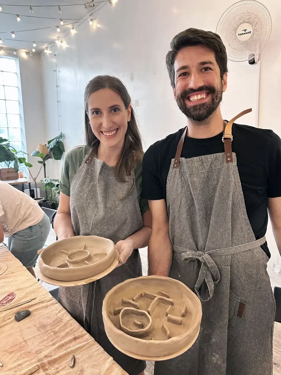 A couple in aprons holding the handmade clay pet bowls they built at a pottery class at Bat Haus in Brooklyn.