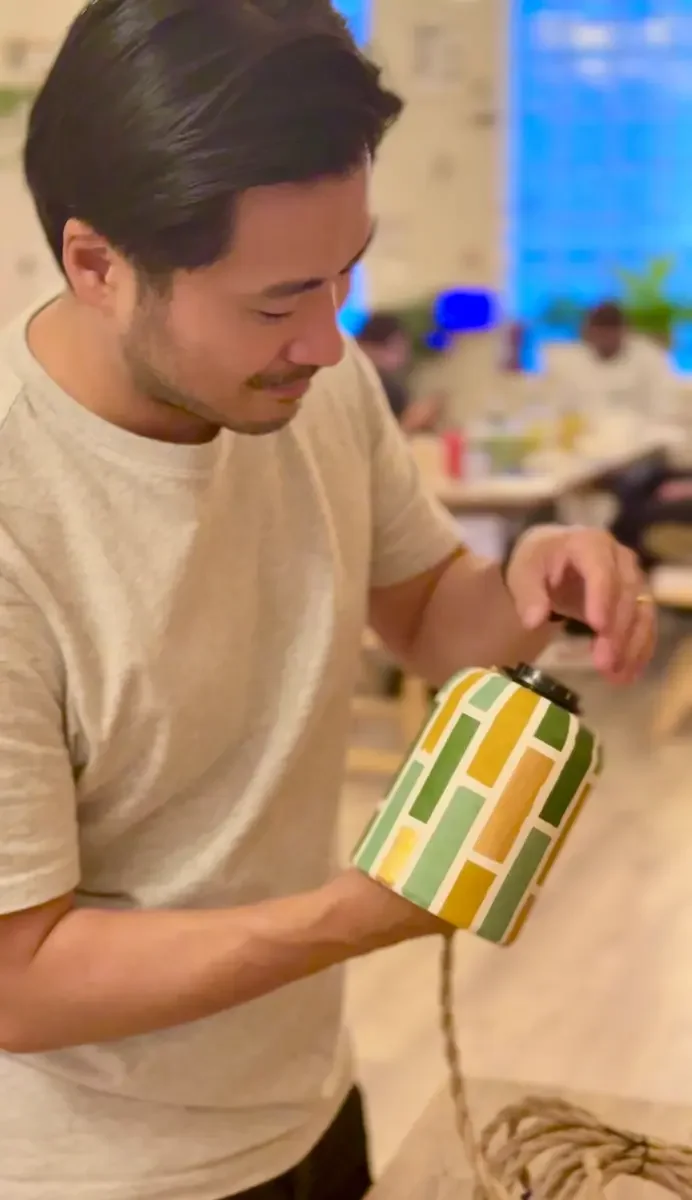 A guest holding a finished green and gold patterned lamp base while attaching the cord and fitting.