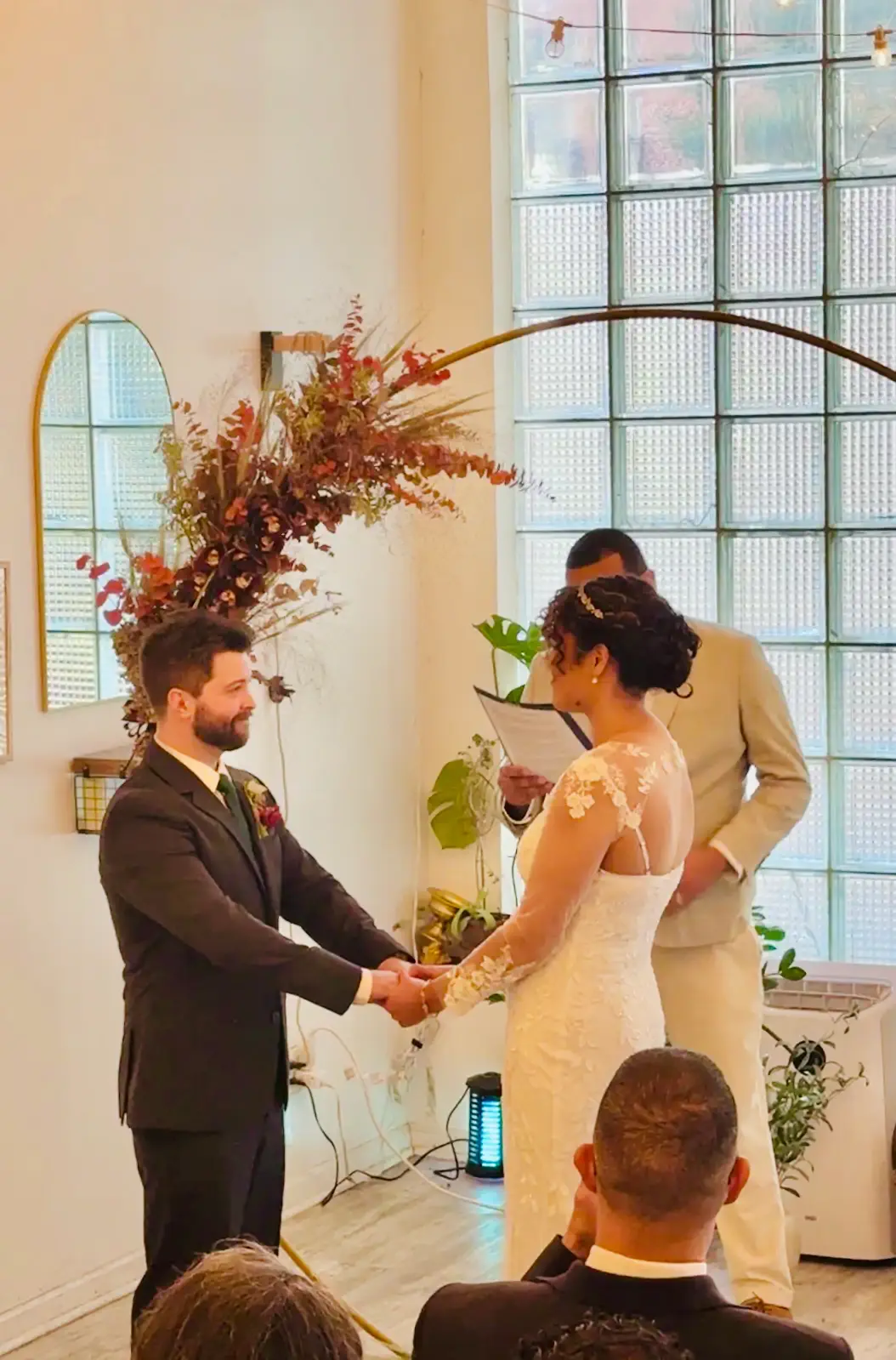 Intimate wedding ceremony at Bat Haus in Williamsburg Brooklyn with bride and groom exchanging vows under a floral arch in a light-filled event space with glass block windows.