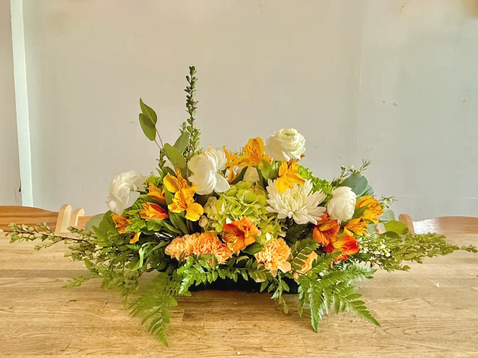 Low and long floral centerpiece with white ranunculus, green hydrangea, orange alstroemeria, and fern greenery designed for Bat Haus wooden tables in Brooklyn event space.