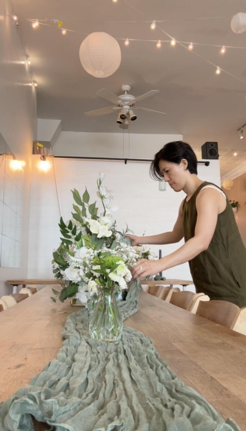 Host arranging white floral centerpieces on a long wooden table with a sage green runner during Nikah setup at Bat Haus in Williamsburg, Brooklyn