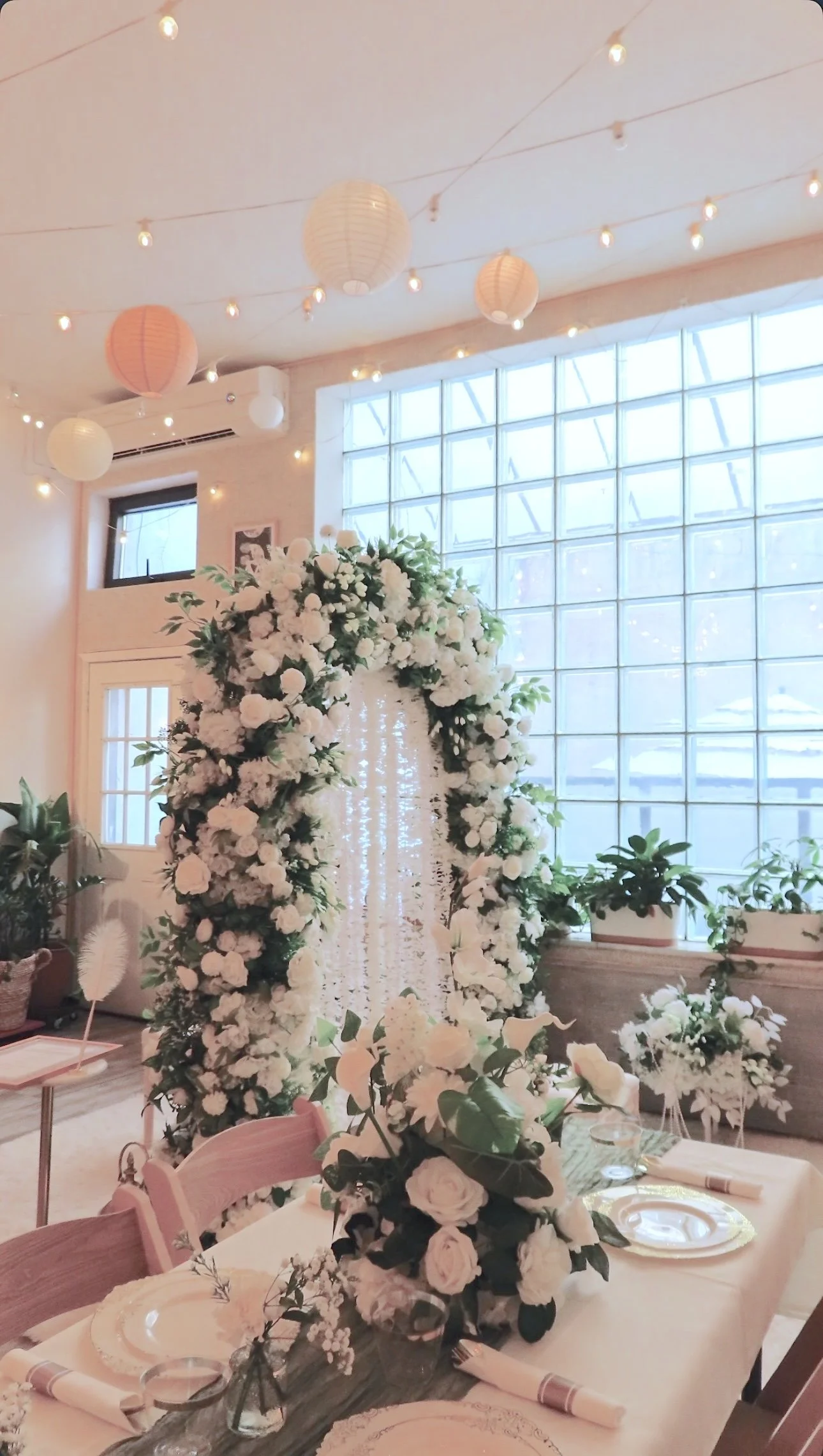 White floral ceremony arch with hanging garlands in front of the glass-block window at Bat Haus in Williamsburg, Brooklyn, with reception tables set in white linens and gold-rimmed plates for a Nikah celebration