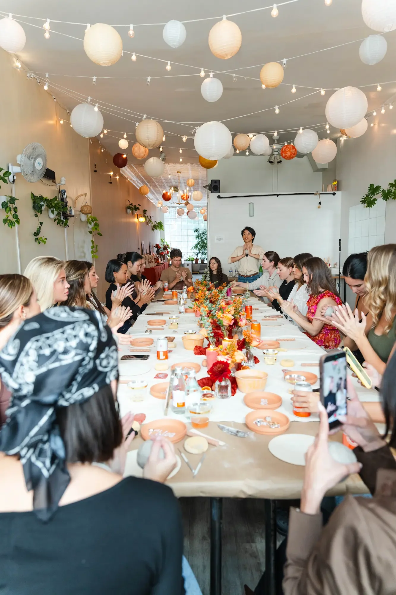 About 20 guests at a group pottery class around one long table at Bat Haus event space in Brooklyn, under paper lanterns and string lights.