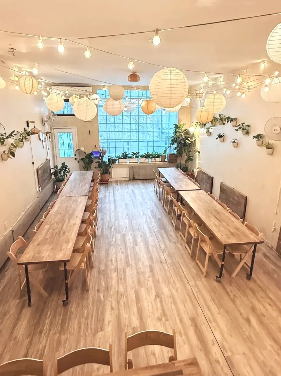 Main seating area at Bat Haus Brooklyn event space featuring five long wooden banquet tables for 40 to 50 guests, string lights, paper lanterns, and greenery along the walls.