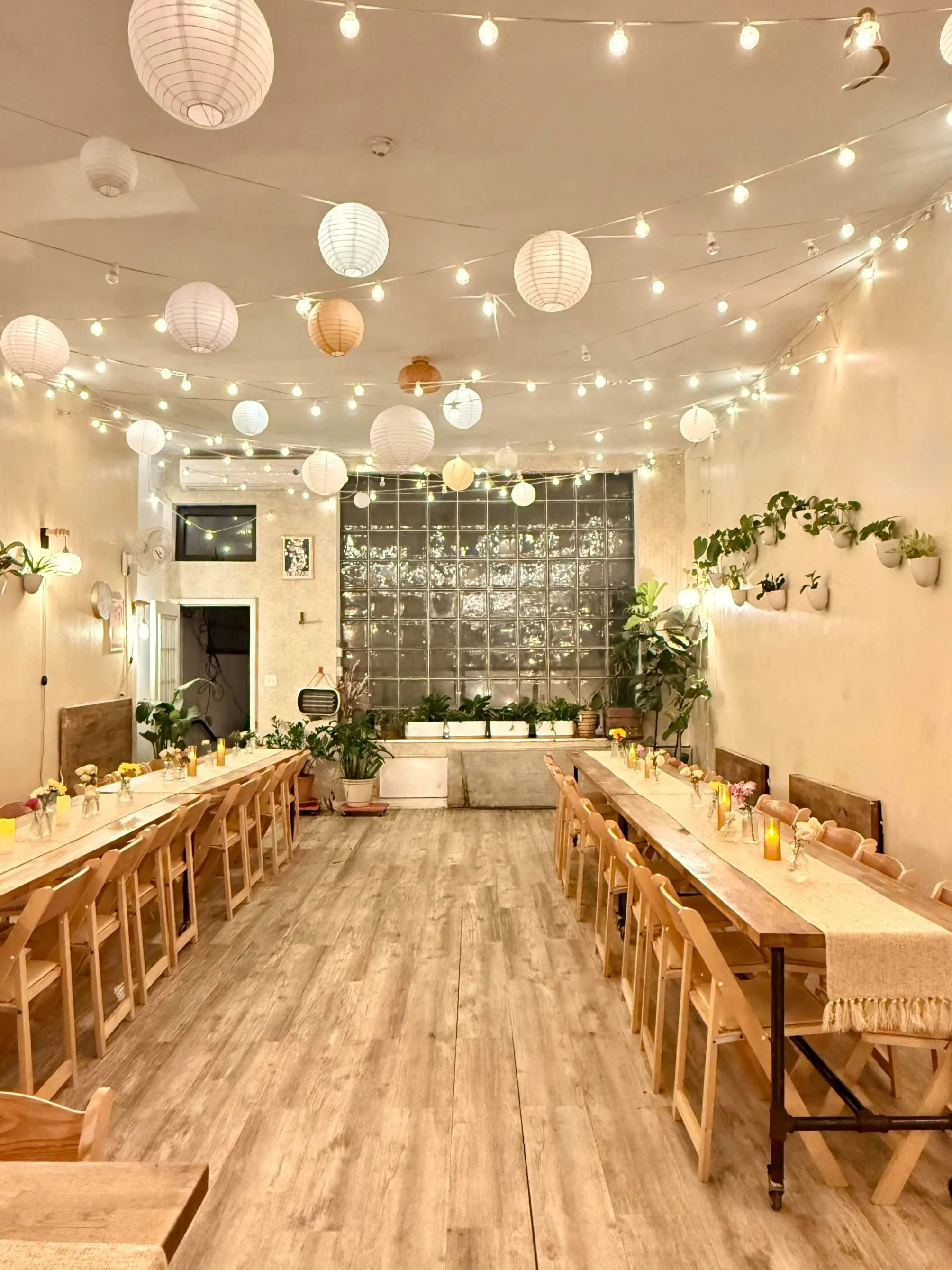 Intimate wedding reception setup at Bat Haus in Brooklyn featuring long wooden tables, warm string lights, paper lanterns, greenery, and a cozy, light-filled atmosphere.