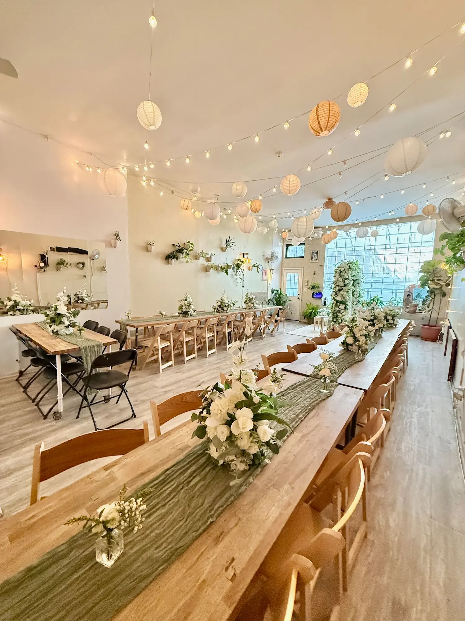 Full view of Bat Haus in Williamsburg, Brooklyn set up for a 50-guest Nikah — long wooden tables with sage green runners and white florals, a floral ceremony arch by the glass-block window, and paper lanterns overhead