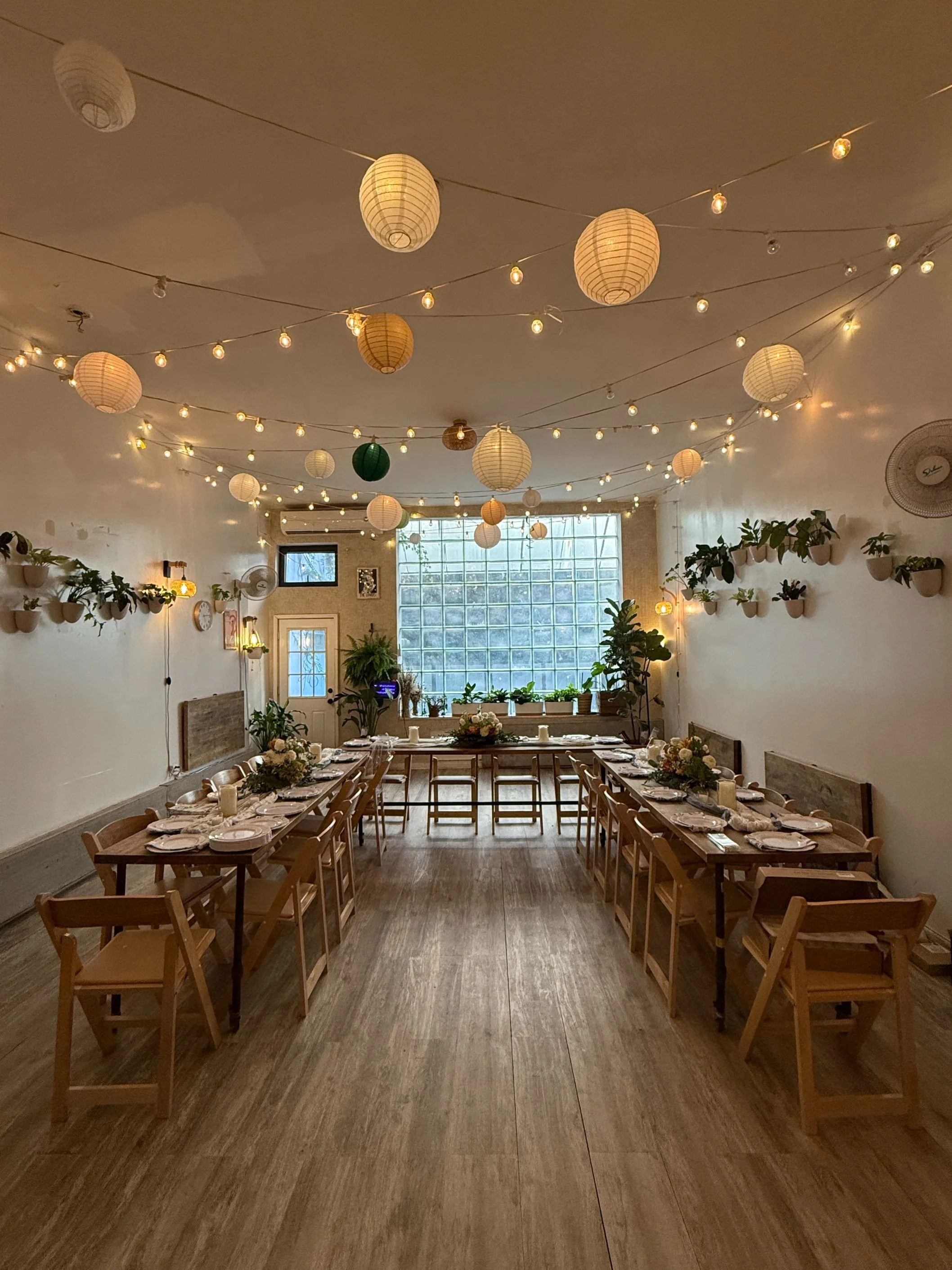Main room set for an intimate dinner: three 8×2′ wooden tables in a U-shape with our low-long centerpieces down the middle. Warm string lights and paper lanterns glow overhead, wall-hung plants frame the room, and the glass-block window anchors the v