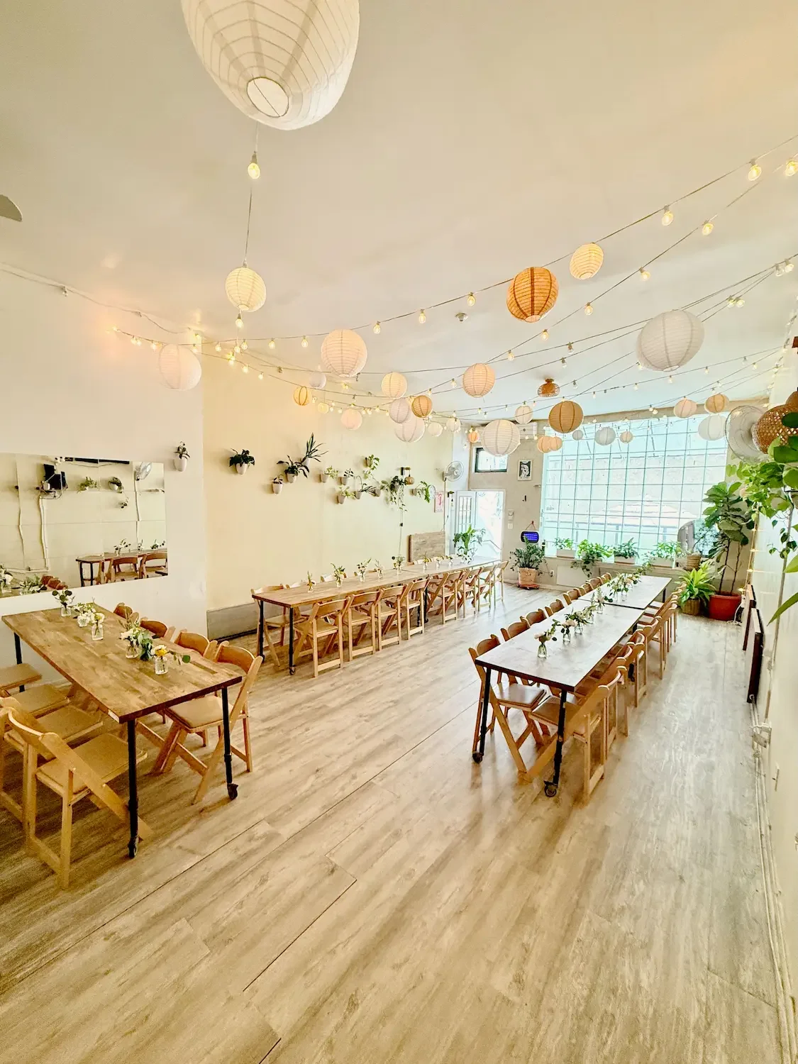 Bat Haus main room in Williamsburg, Brooklyn set with long wooden farm tables, folding chairs and bud-vase florals under paper lanterns and string lights, with a plant-lined wall and large windows of natural light.