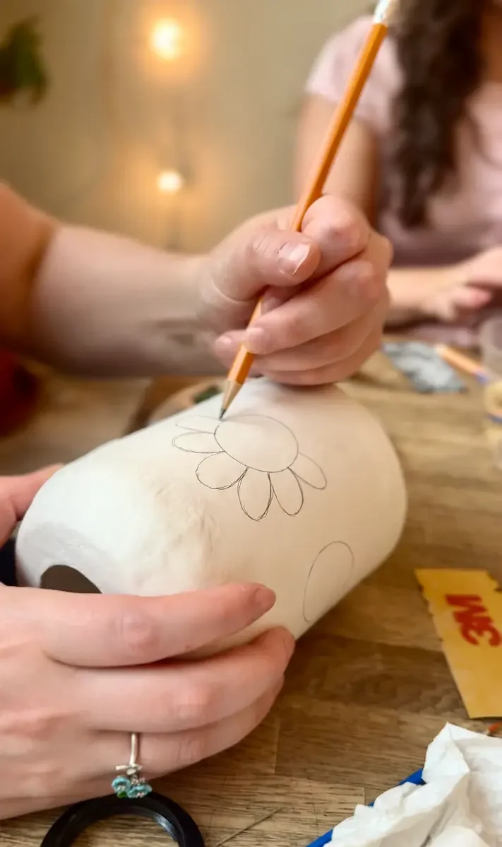 Hands sketching a flower design in pencil onto a white ceramic lamp base at a lamp making workshop.