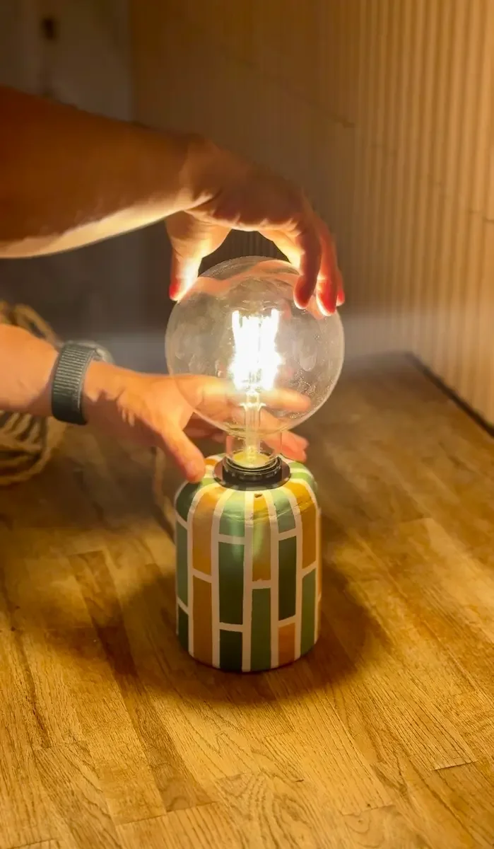Hands placing a glowing Edison bulb into a finished hand-painted lamp base on a wooden table.