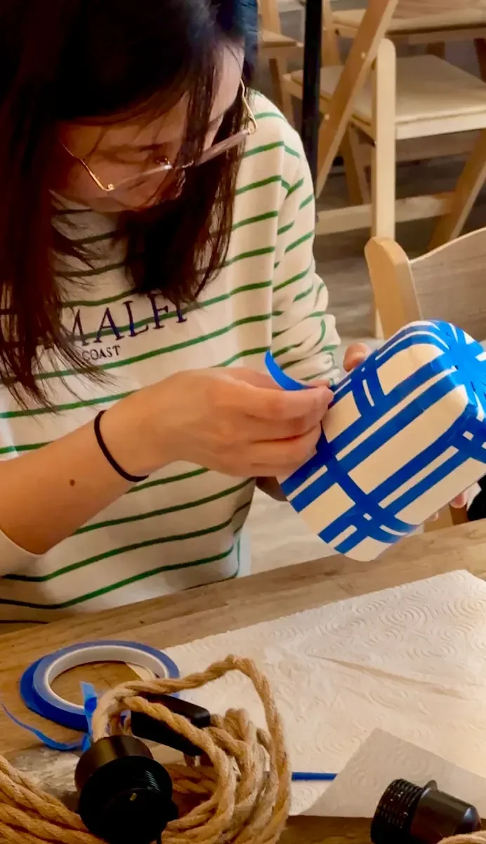 A workshop guest using blue tape to create a plaid pattern across a ceramic lamp base.