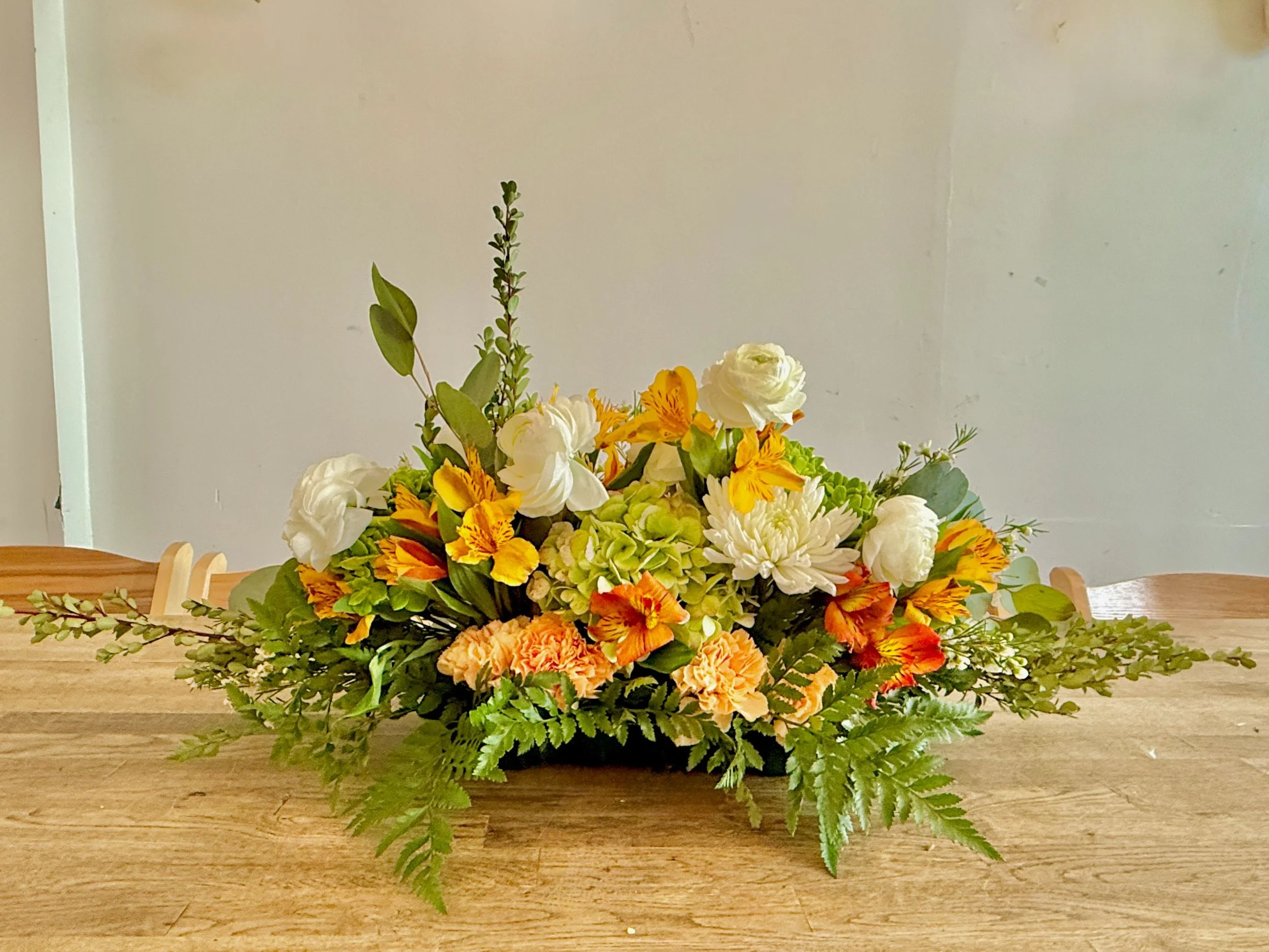 Citrus Meadow — Cream & Marigold
A wide, low centerpiece with cream ranunculus and mums, fresh green hydrangea, and sunlit marigold/tangerine alstroemeria. Fern and eucalyptus soften the edges for an easy, conversation-friendly profile.
Vibe: bright