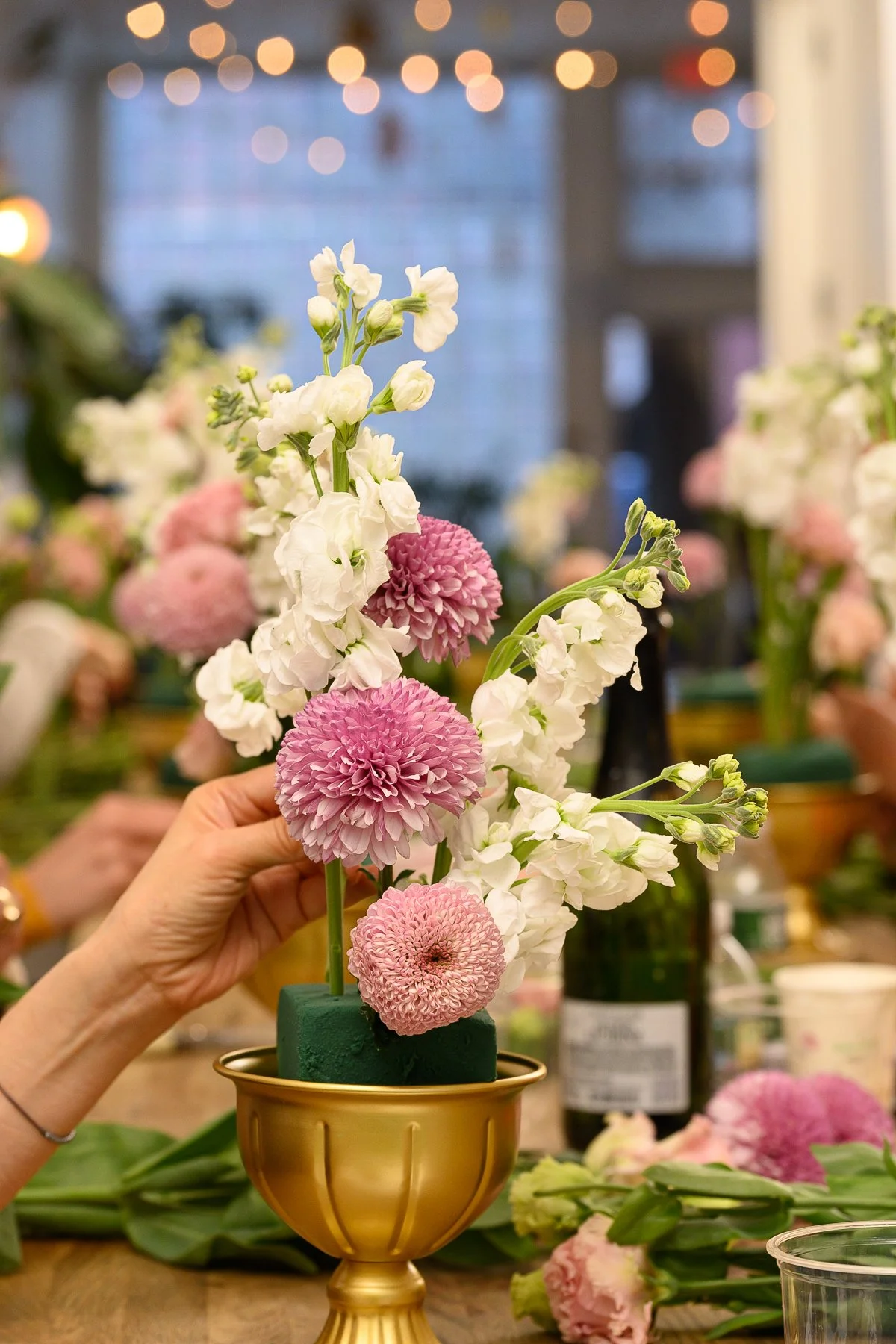 Hands arranging pink and white blooms into a gold pedestal vase at a bridal floral workshop at Bat Haus in Williamsburg, Brooklyn.