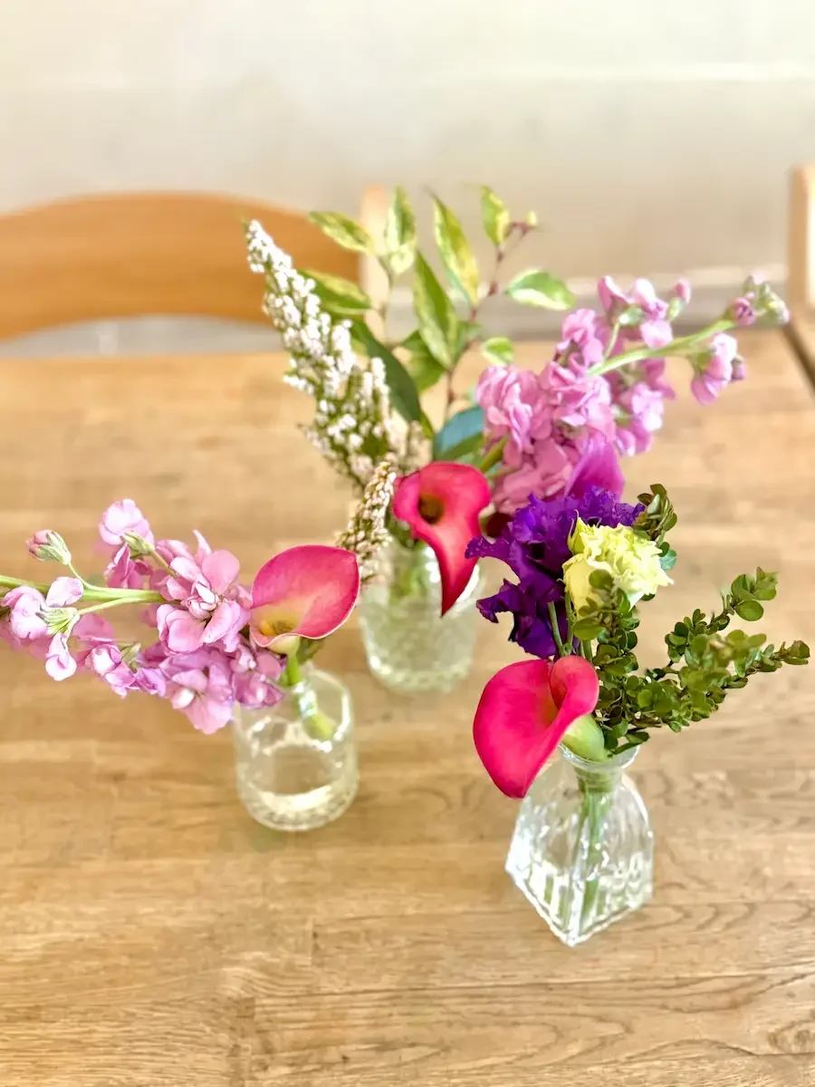 Pink and purple bud vase table florals with calla lilies and stock on a long wood table at Bat Haus in Williamsburg, Brooklyn.