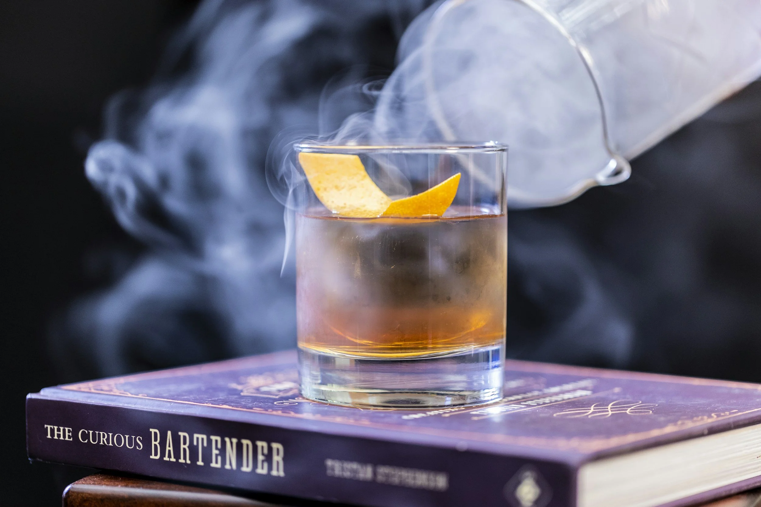 A glass of cocktail with a lemon garnish, placed on top of a book titled 'The Curious Bartender', with smoke or vapor in the background.