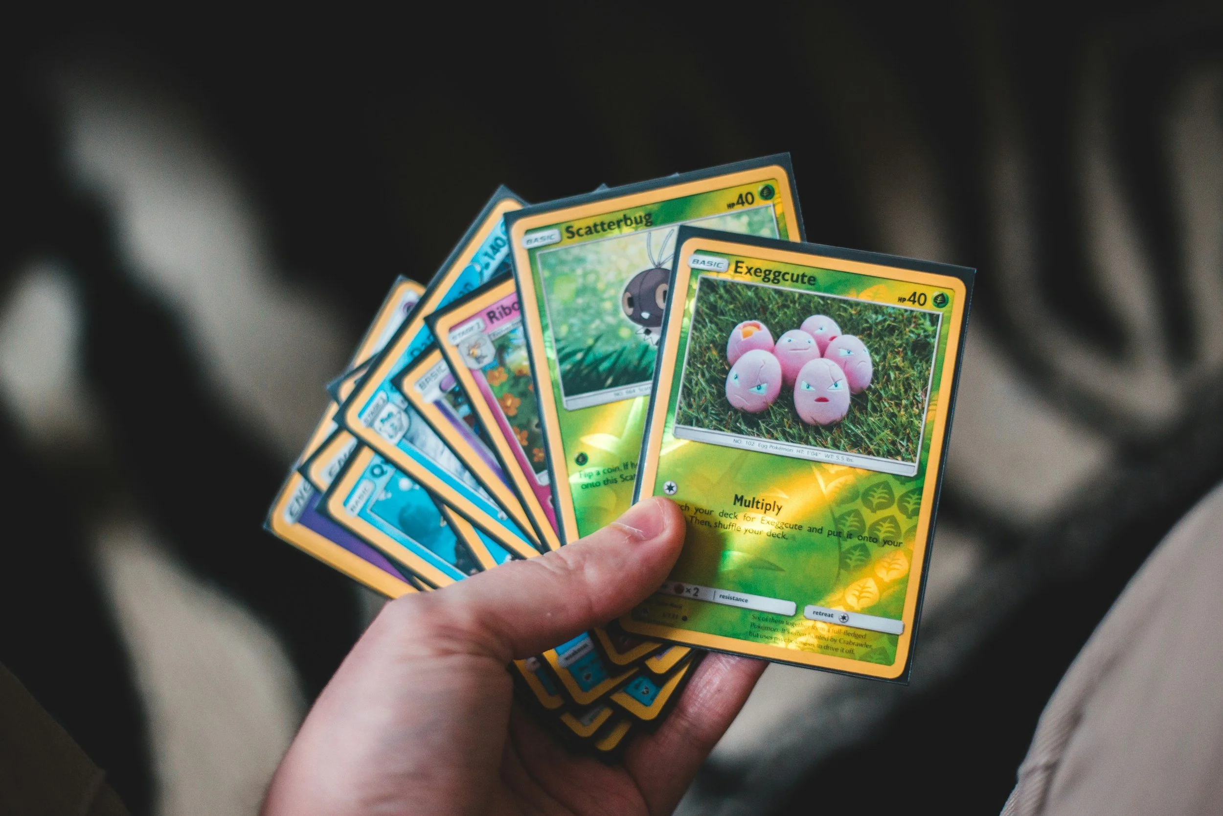 A hand holding several Pokémon trading cards fanned out, with the first two cards showing the Pokémon Exeggcute and Scatterbug.