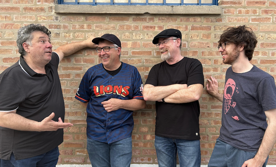 Four men standing outside against a brick wall, engaged in conversation, smiling and gesturing.