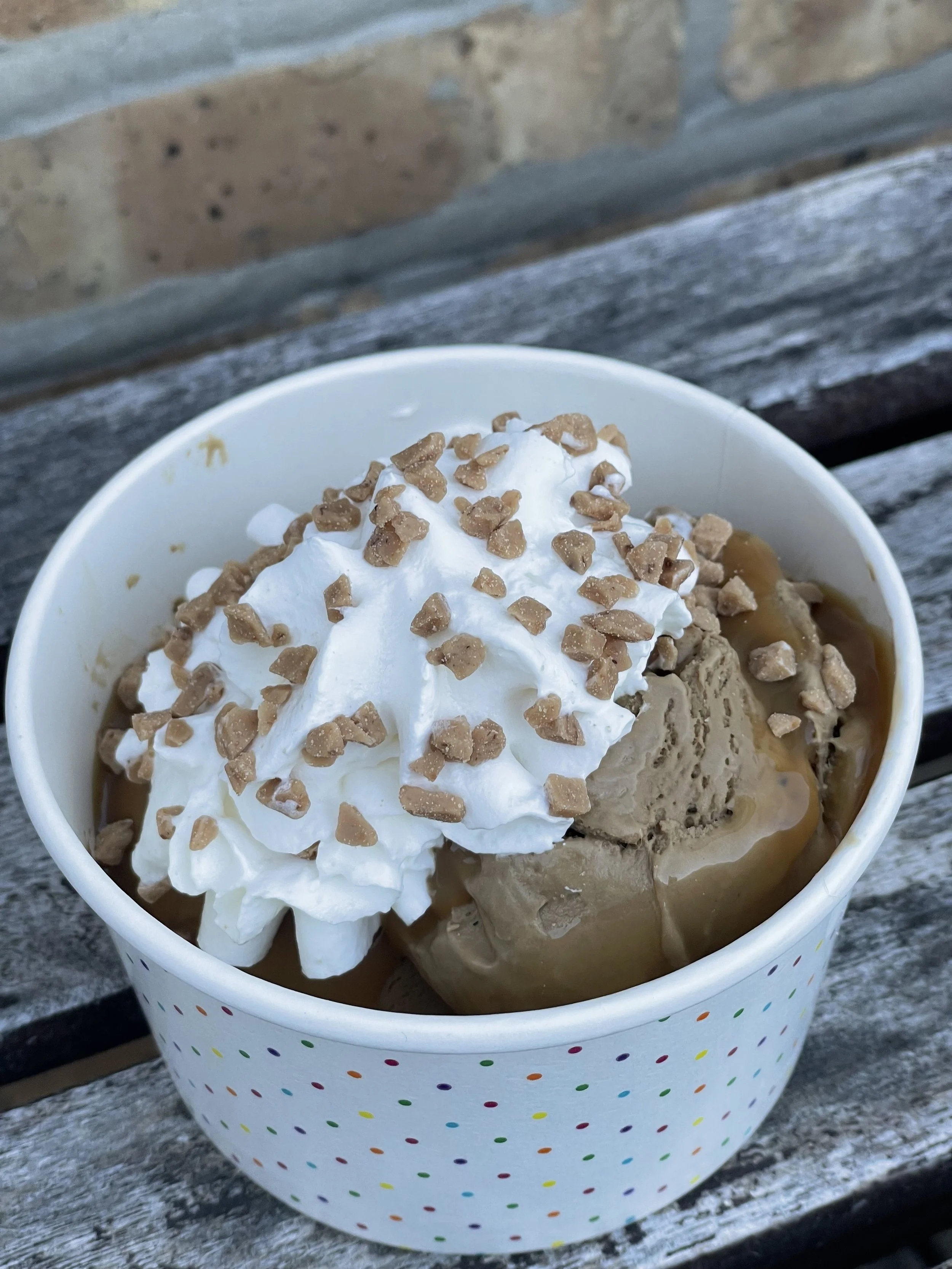 Ice cream with whipped cream and chopped nuts in a polka dot paper cup on a wooden surface.