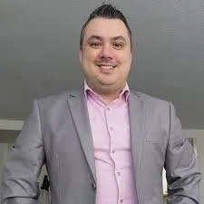 Smiling man in a gray suit jacket with a pink shirt standing indoors