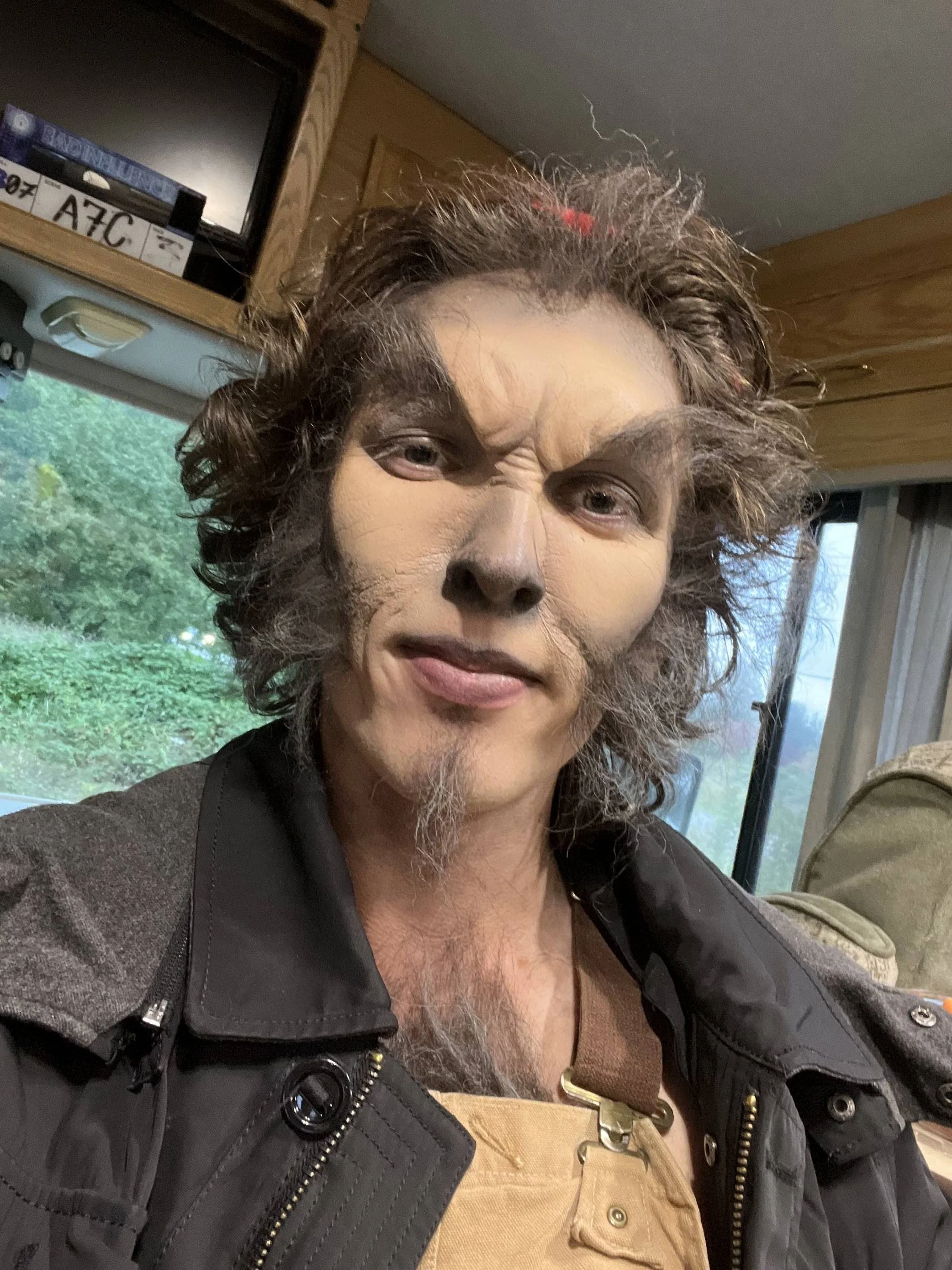 A person with makeup and prosthetics that give a hairy, scruffy appearance, with wild hair and a beard, wearing a black jacket and beige work overalls, inside a camper or RV with greenery visible outside the window.