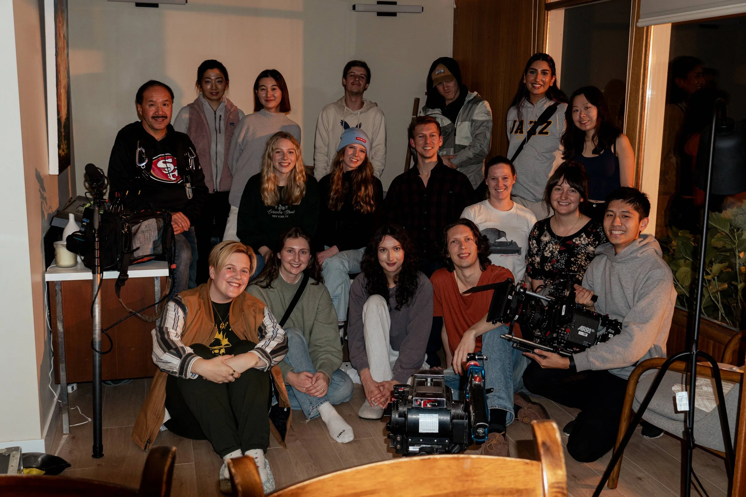 A group of 17 young adults in a cozy indoor space, posing for a photo, with some filming equipment visible in the foreground.