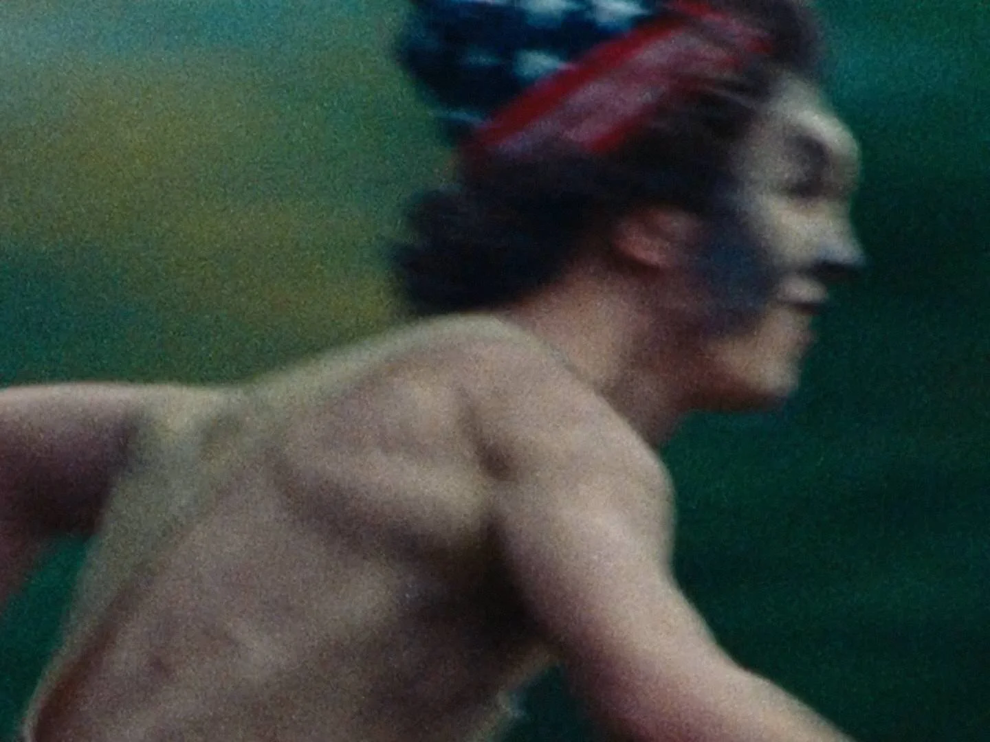 A topless young person with dark, three-colored hair styled in a voluminous way, smiling, against a green background.