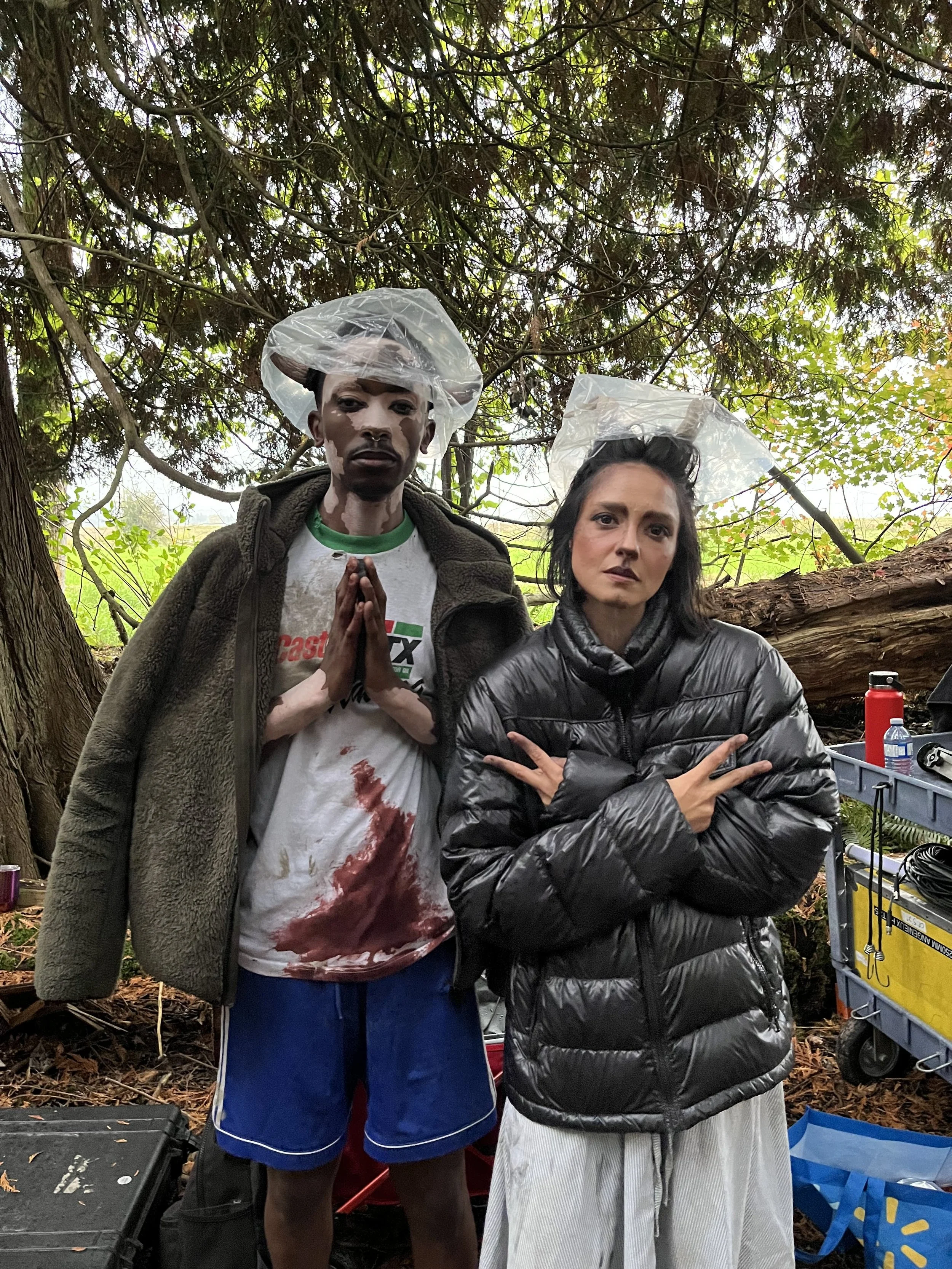 Two people standing outdoors in a wooded area, both wearing transparent plastic face shields. The person on the left has dark skin, a beard, and is wearing a gray fur-lined jacket over a white t-shirt with bloodstains and a green collar, and blue sho
